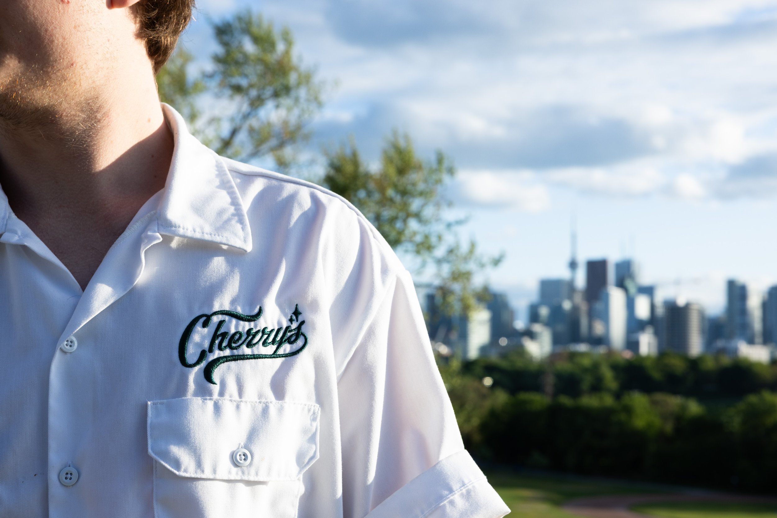 A person wearing a white shirt with the embroidered name "Choroll's" standing outdoors with a city skyline in the background.
