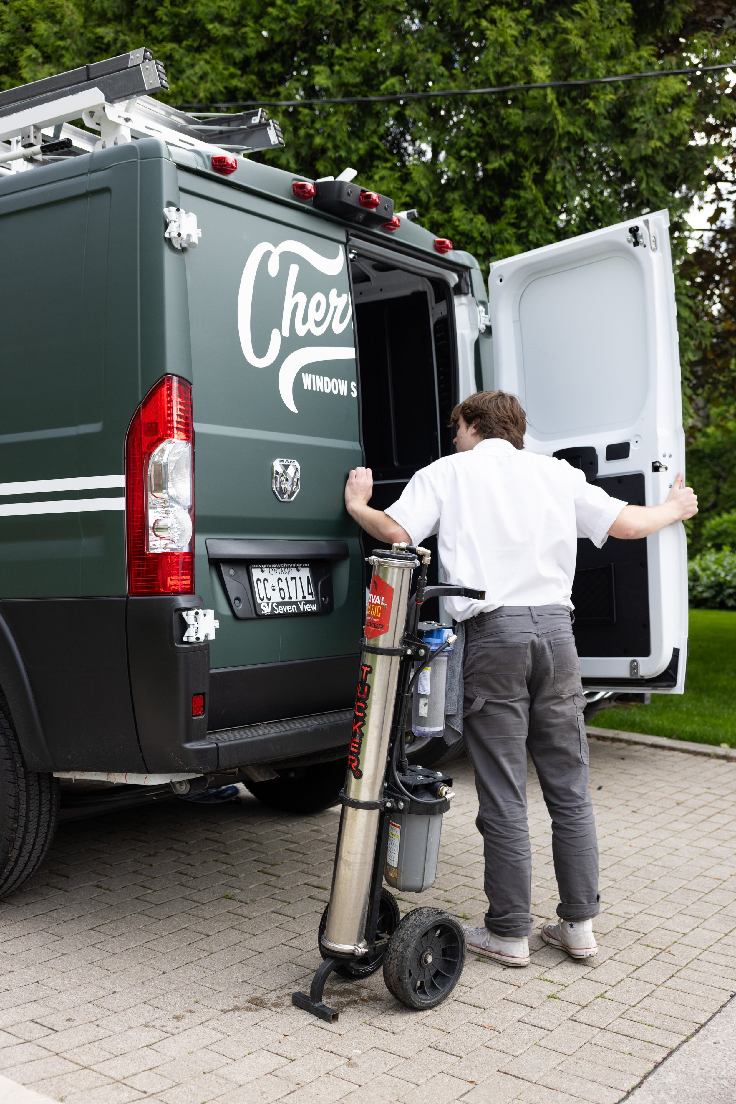 A worker in a white shirt and gray pants with a nerve device, standing by a green window repair van labeled "Cher" and "Window". The van's rear door is open, and the worker is preparing to service the van's interior. The van is parked on a brick driv