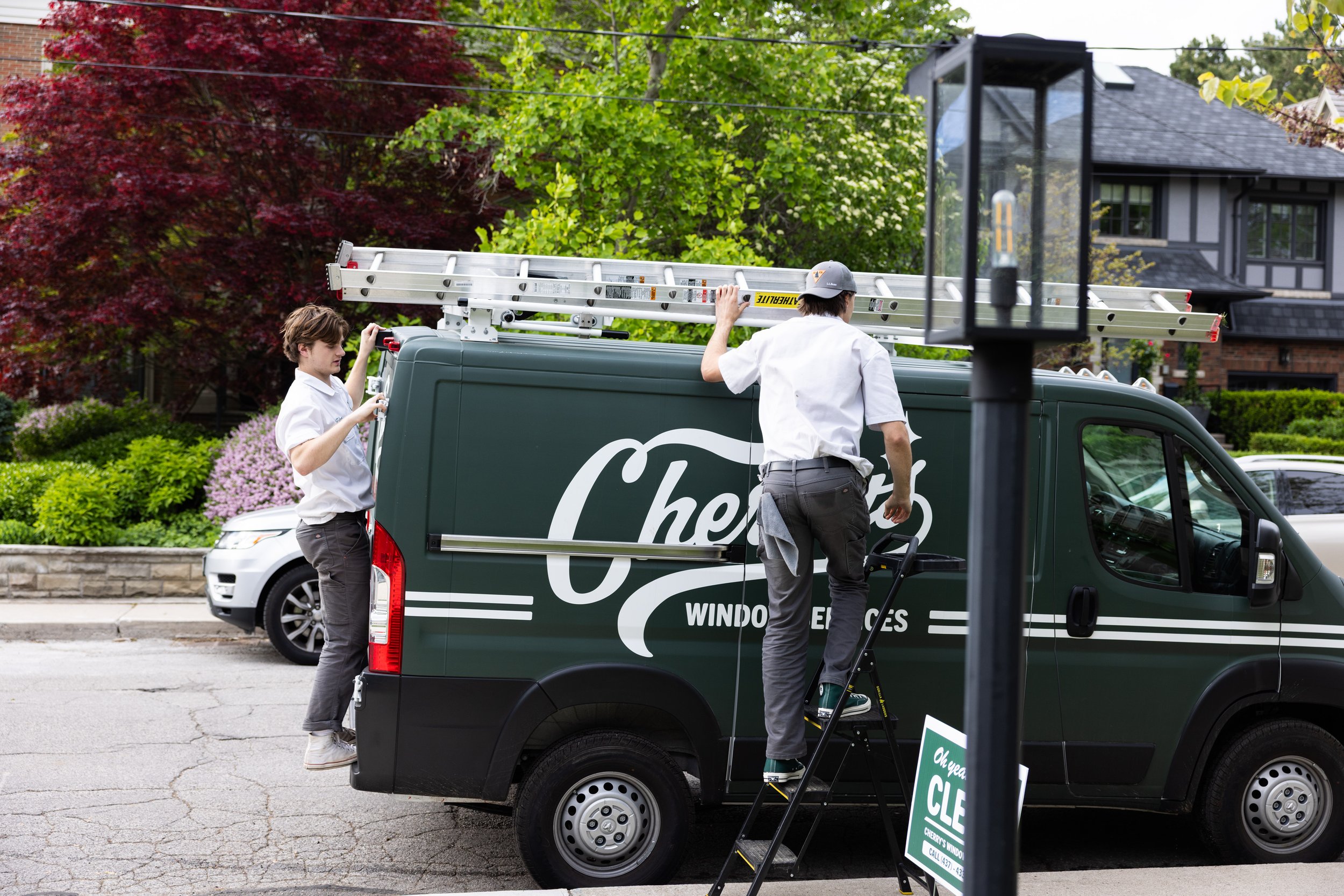 Two workers in white shirts and gray pants installing a sign on a green window service van, with one stepping on a small ladder and other holding the side of the van, on a residential street with colorful bushes and trees in the background.