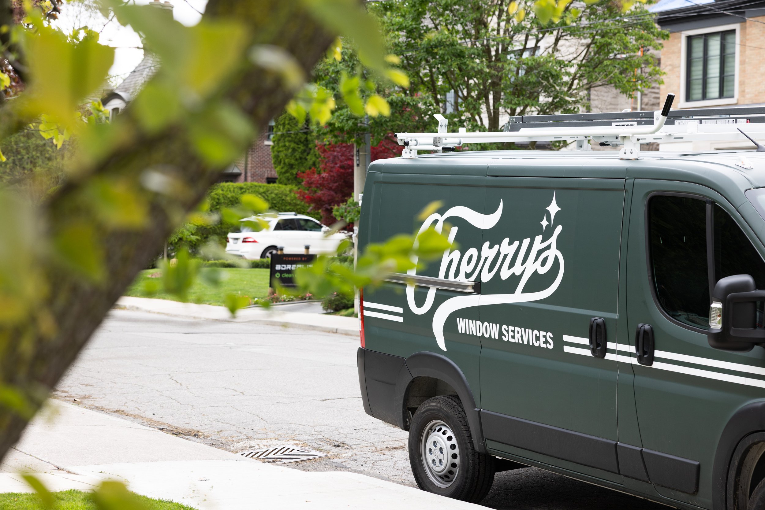 A green window service van with the word "Sherry's" and "Window Services" written on it, parked on a residential street with trees and houses, partially obscured by blurred tree branches in the foreground.