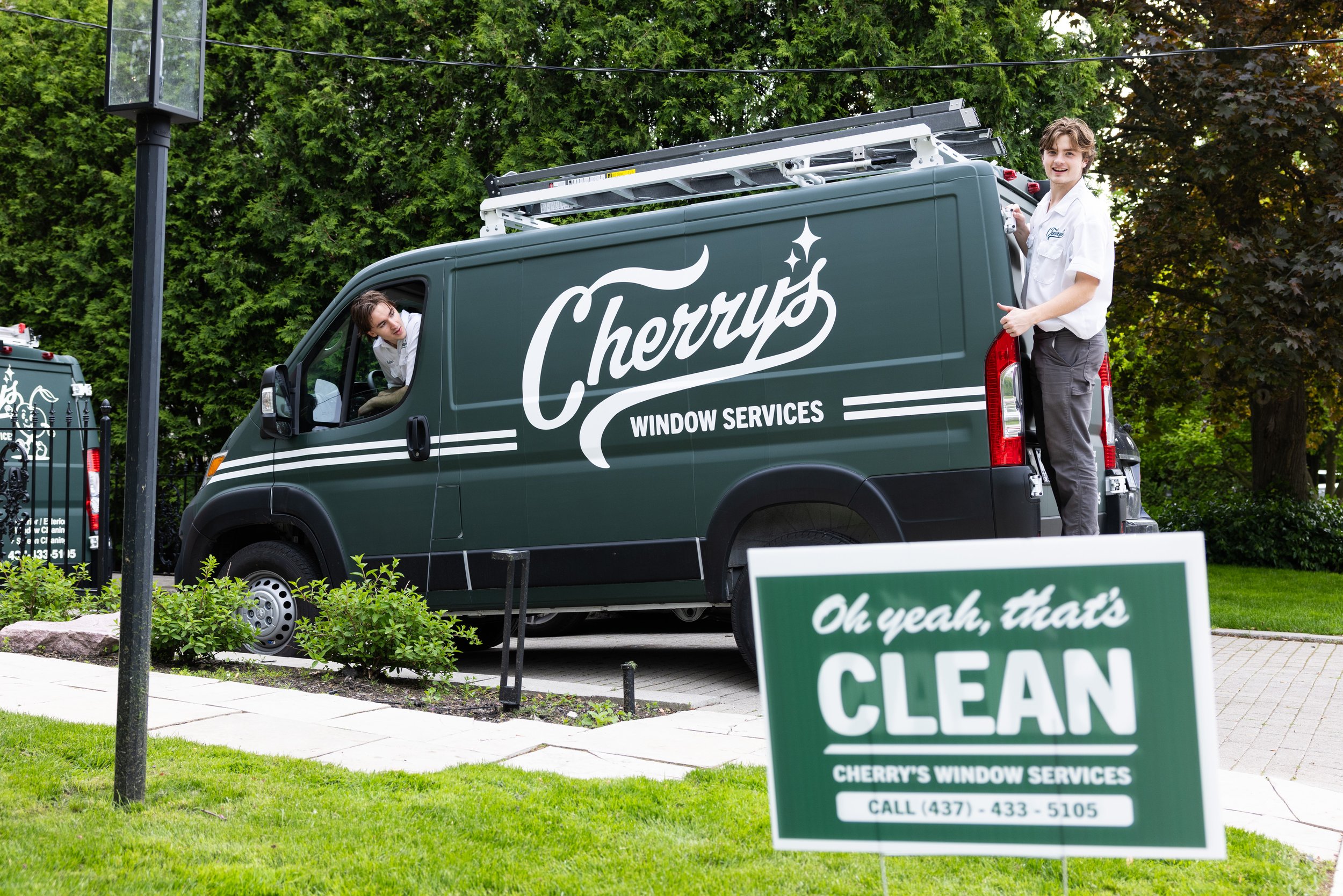A green Cherry's Window Services truck parked outdoors with a worker standing on the rear step, smiling. Another person leans out from the driver's side window. A sign in front advertises Cherry's Window Services with a phone number, emphasizing that