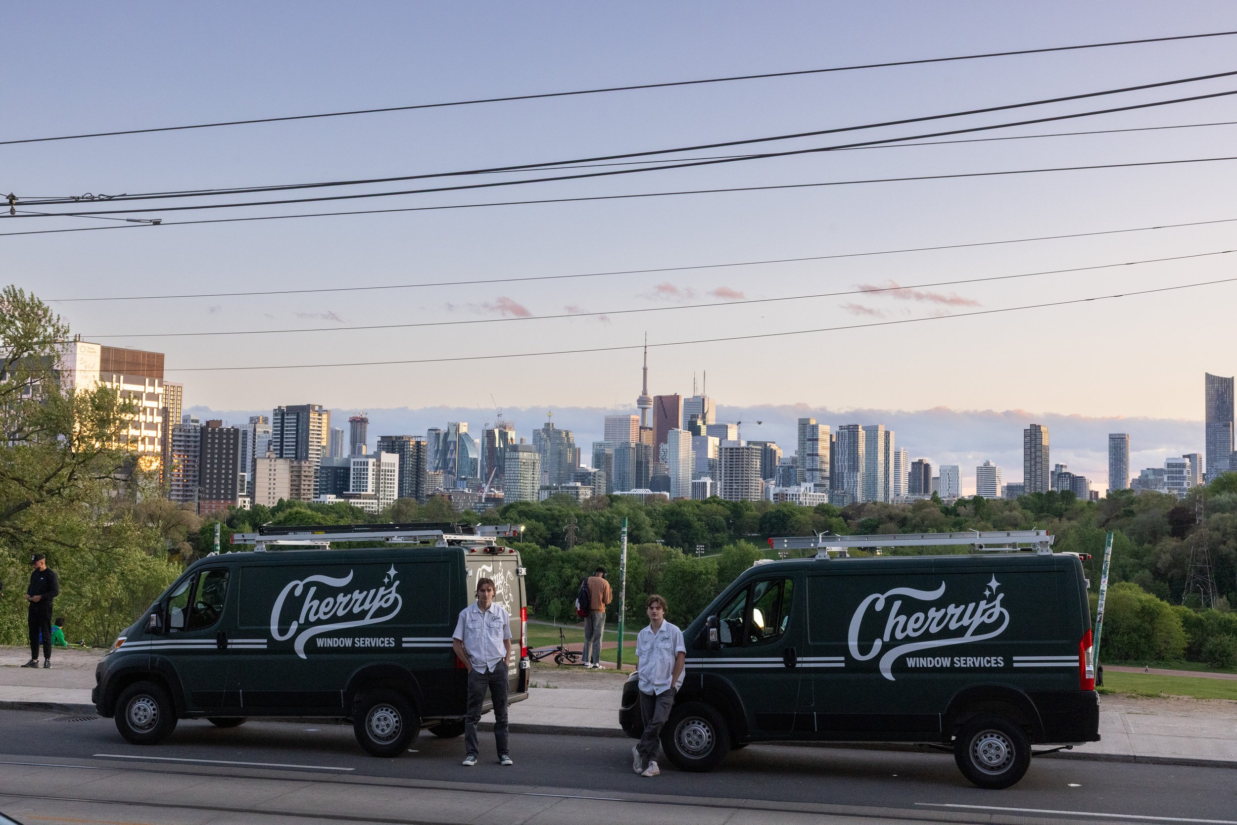 Two Cherry's Window Services vans parked on the street with two workers standing nearby, with a city skyline in the background under a partly cloudy sky.