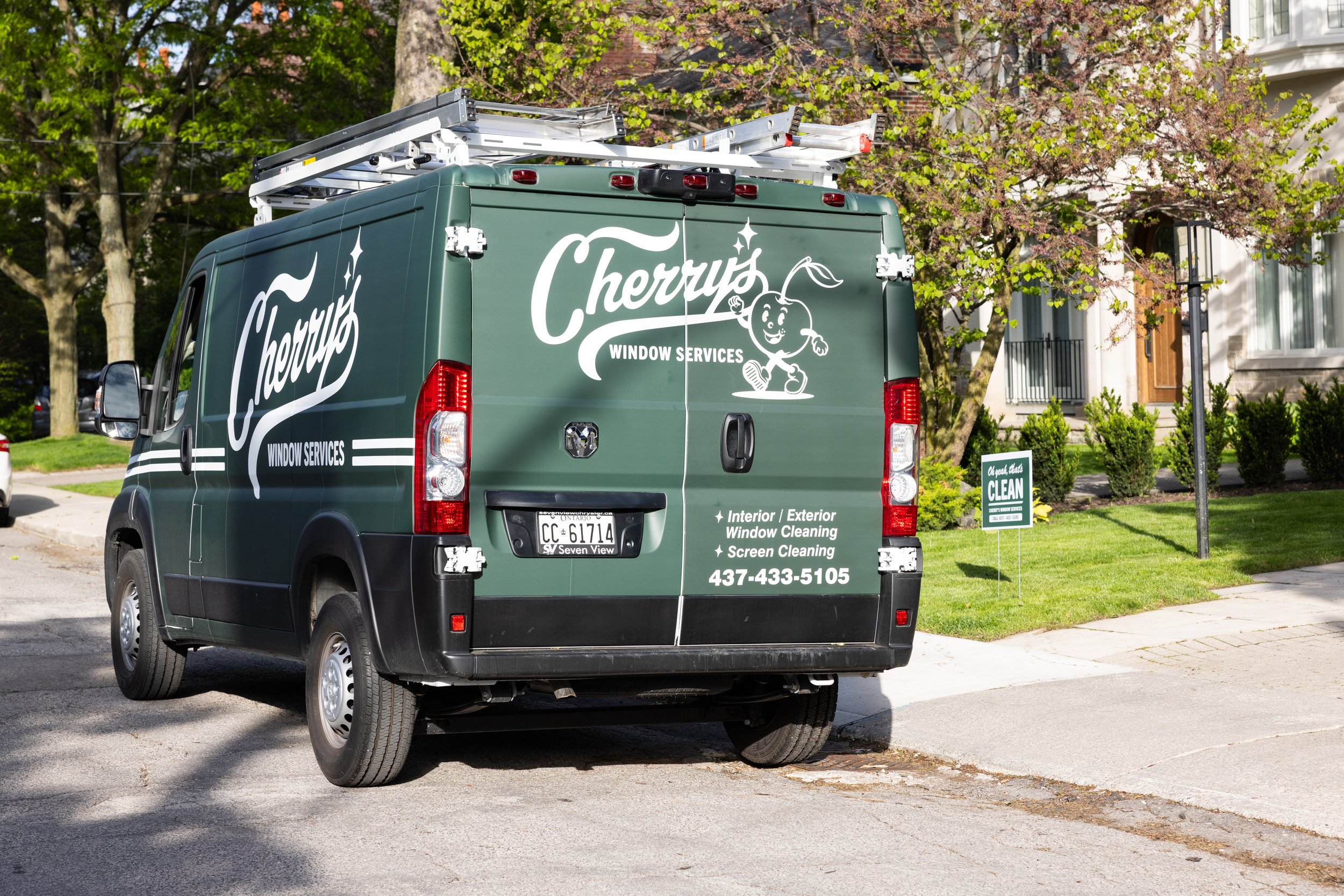 A green Cherry's Window Services company van parked on a residential street with trees and a house in the background. The van has a cartoon cherry character on it and services listed including interior and exterior window cleaning and screen cleaning