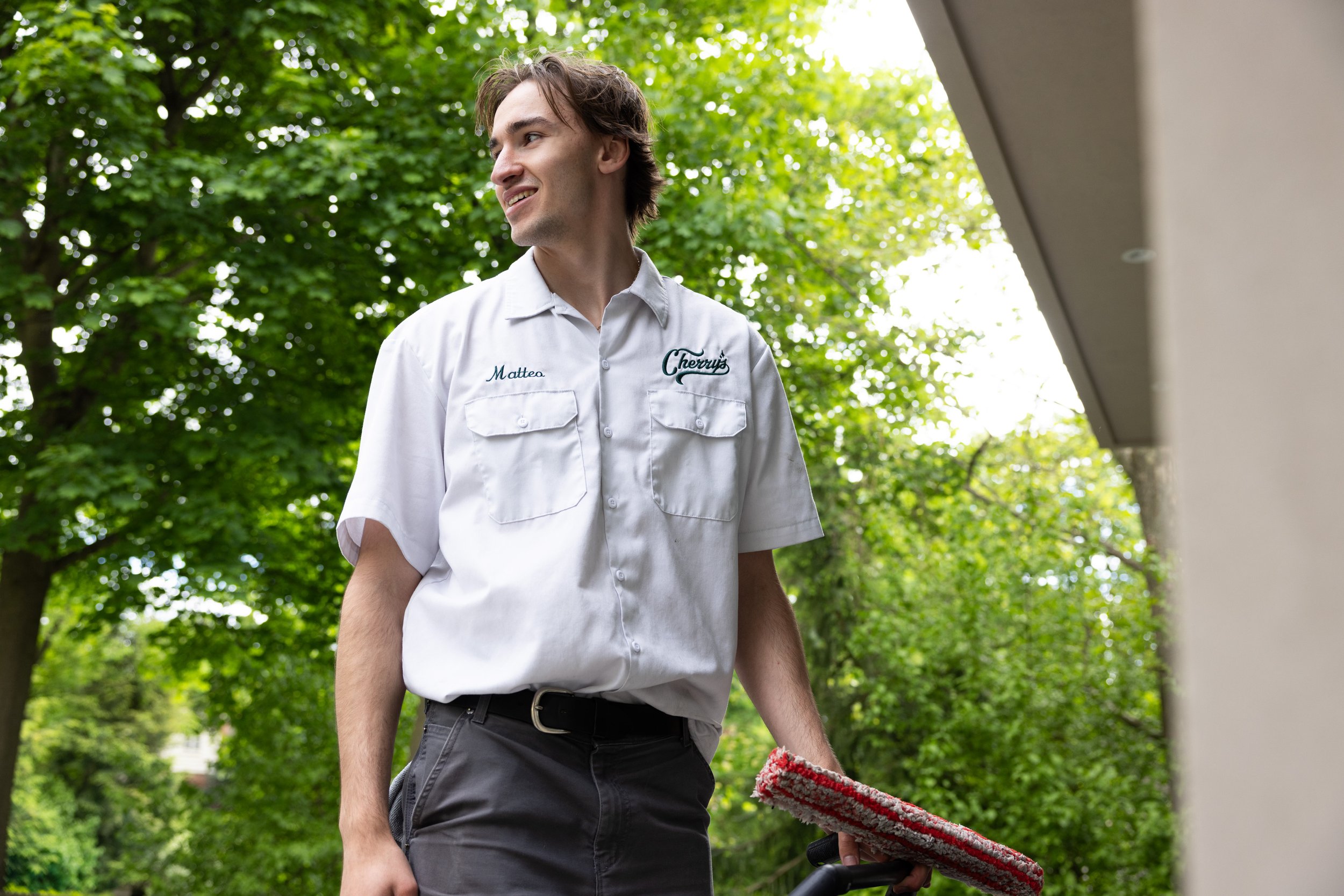 A young man in a white uniform shirt with name tag 'Mattea' and an embroidered logo 'Cherry's' is holding a red and gray scrub brush, standing outside near a building with green trees in the background.