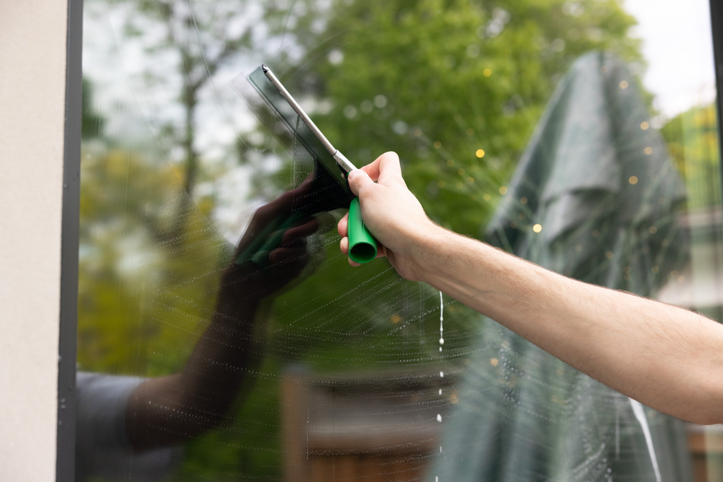 Person cleaning a glass window with a squeegee outside, with green trees reflected on the glass.