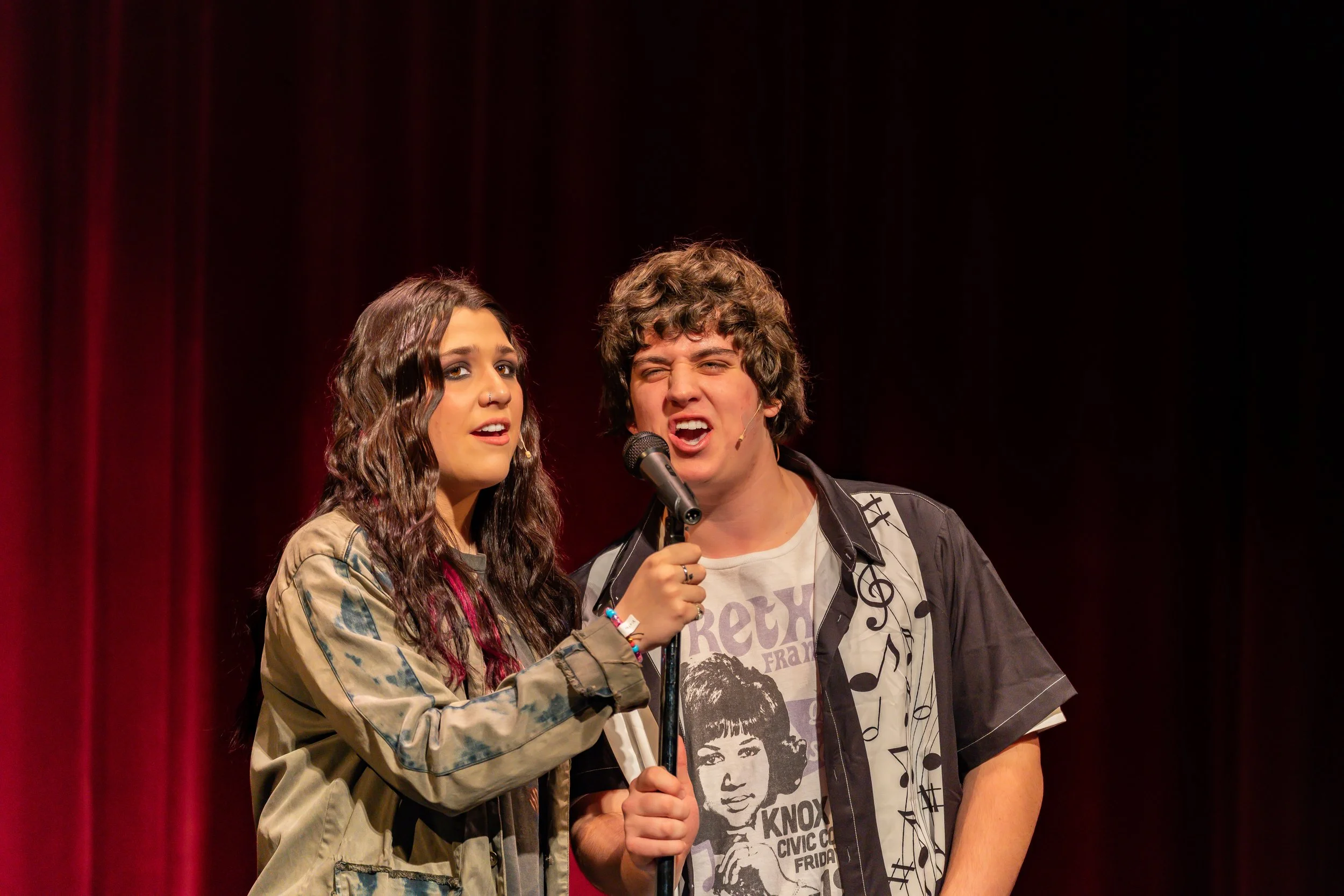 Two young performers singing on stage in front of a red curtain, with one holding a microphone.