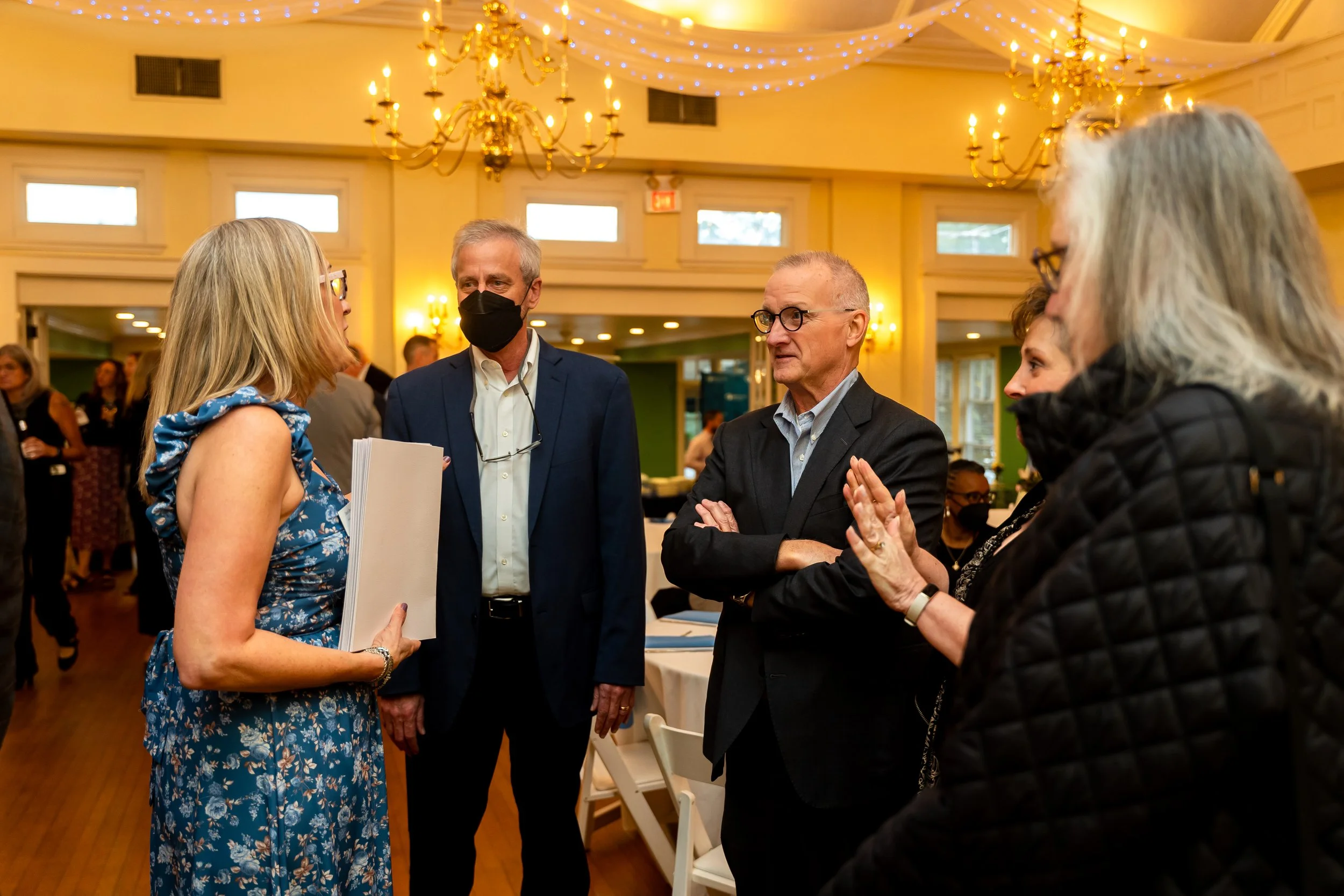 A group of five people, four women and one man, are engaged in conversation at an indoor event with elegant chandeliers and blue string lights. One woman is wearing a blue floral dress and holding a folder, and the man is wearing a navy suit with a b