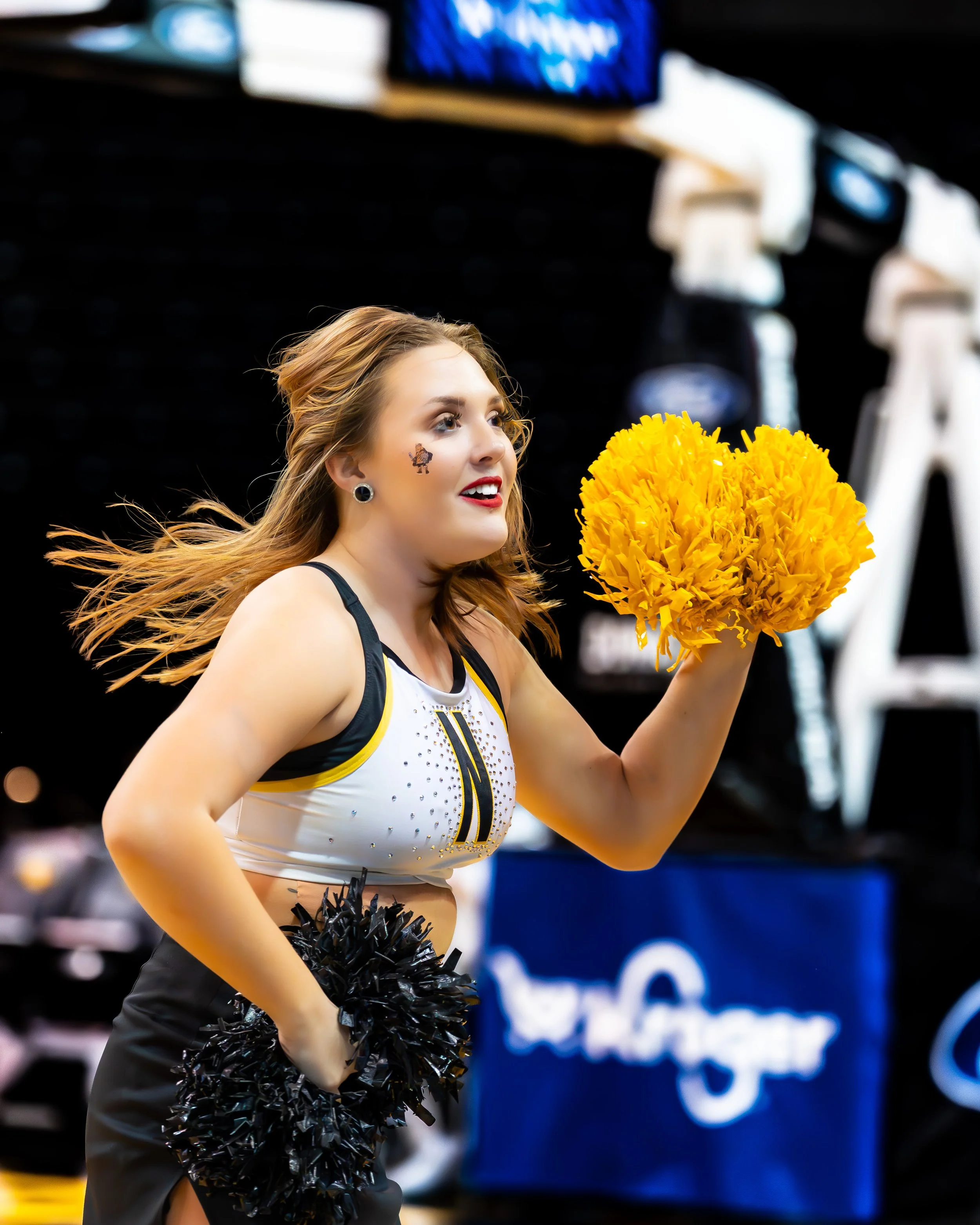 A cheerleader with long, wavy brown hair, wearing a white and black uniform with yellow accents, holding yellow and black pom-poms, smiling at an athletic event.