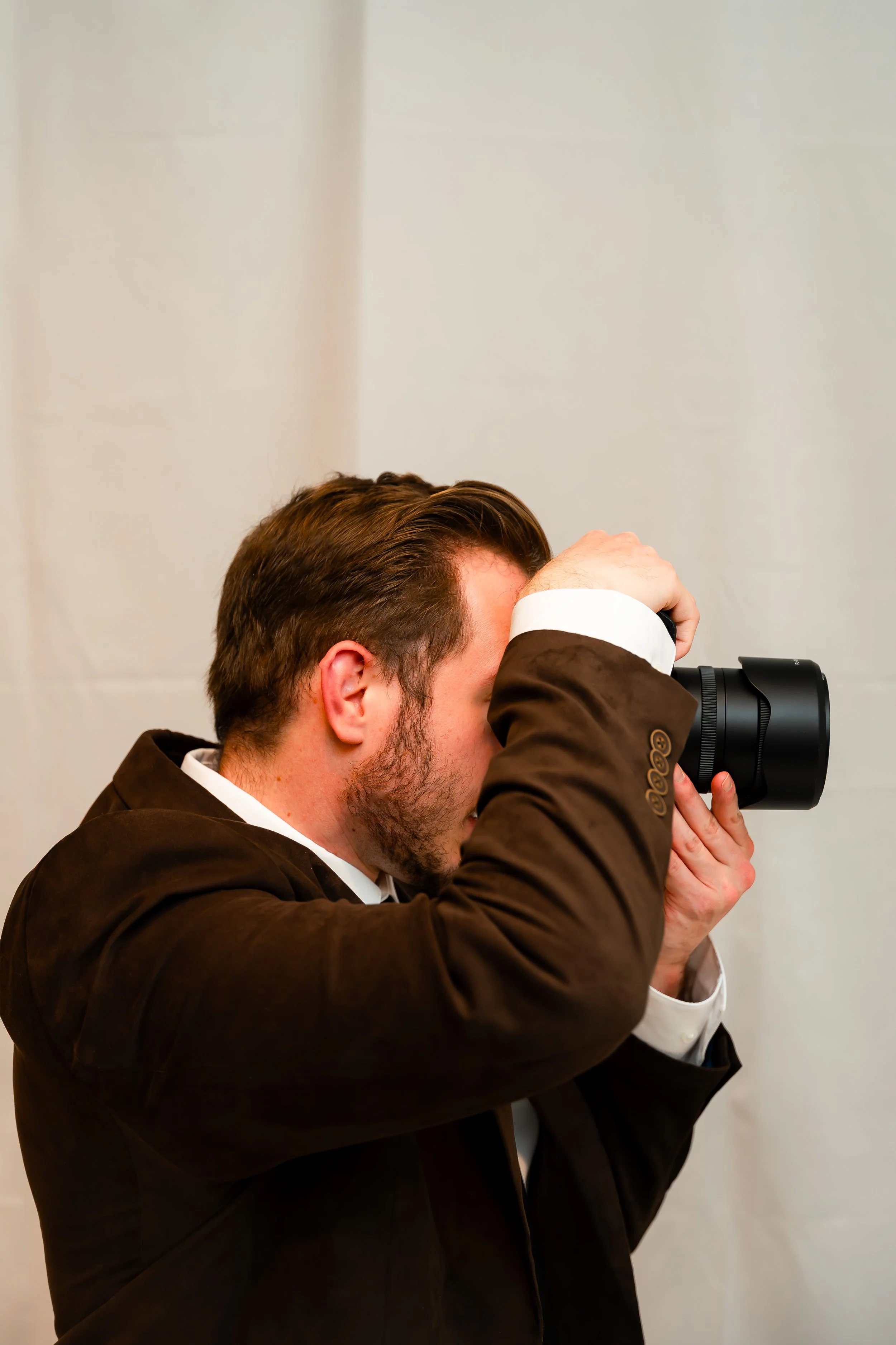 A man with brown hair and beard, dressed in a dark brown blazer and white shirt, is looking through a black camera or binoculars, with his hands holding the device, against a plain light-colored background.
