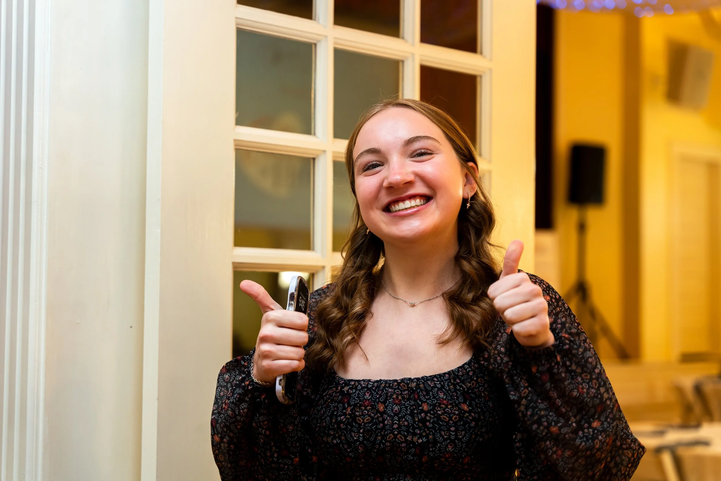 A young woman with long, wavy brown hair smiling and giving a thumbs-up, standing in front of a yellow door with glass panes.