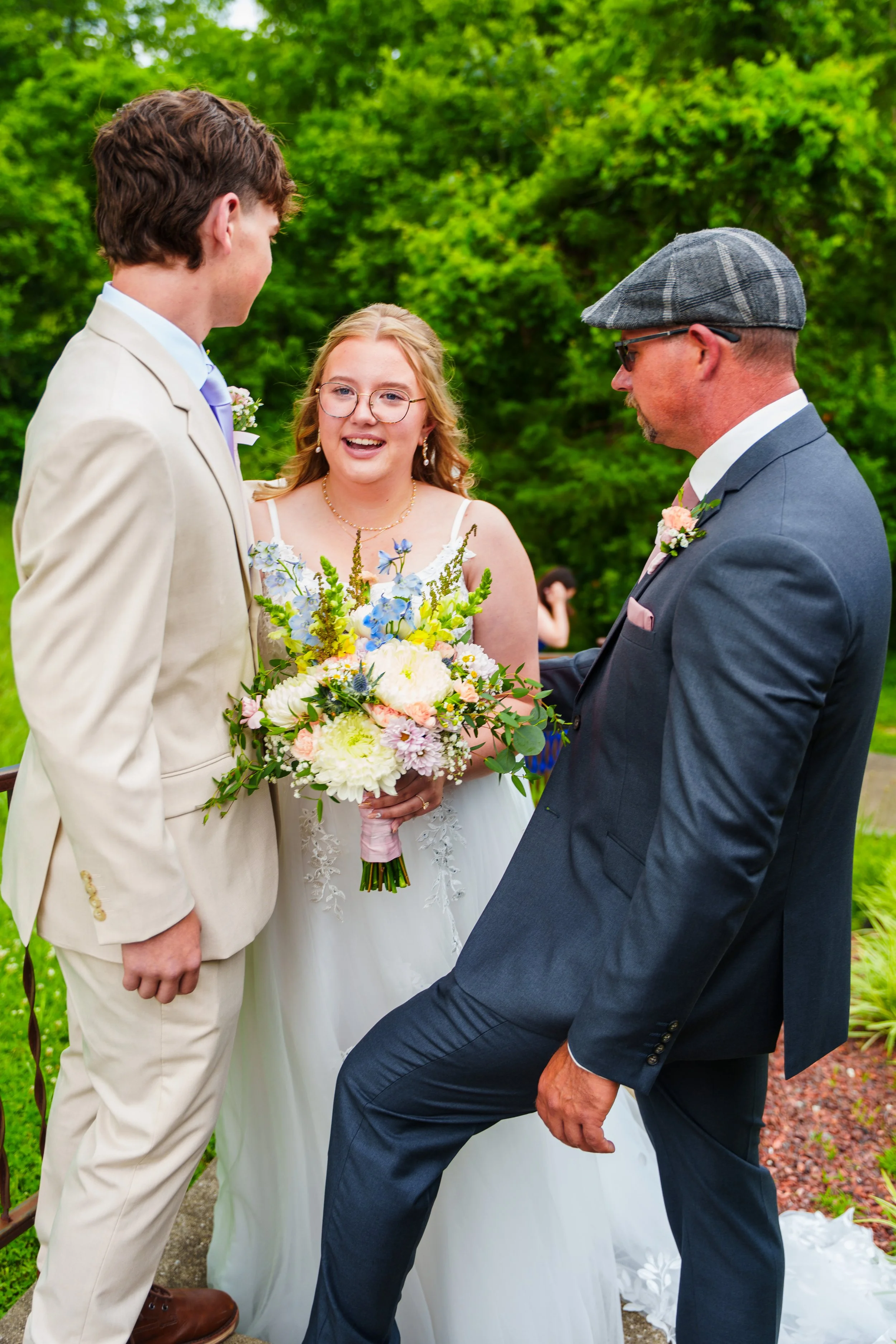 A bride and groom standing outdoors during a wedding ceremony, with the officiant kneeling in front of them, holding their hands, surrounded by greenery.