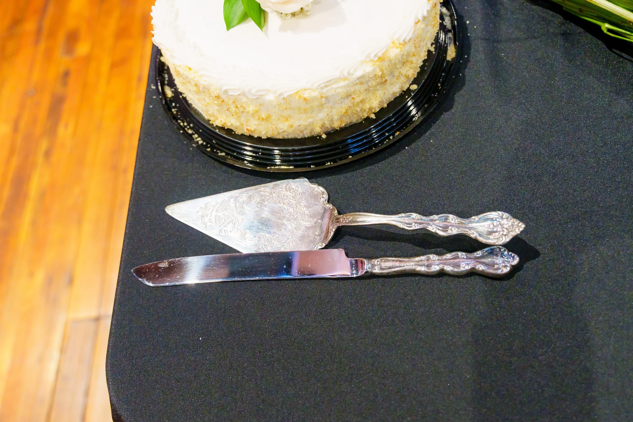 A round white frosted cake on a black plate, placed on a black tablecloth. Silver cake server and knife set next to the cake.