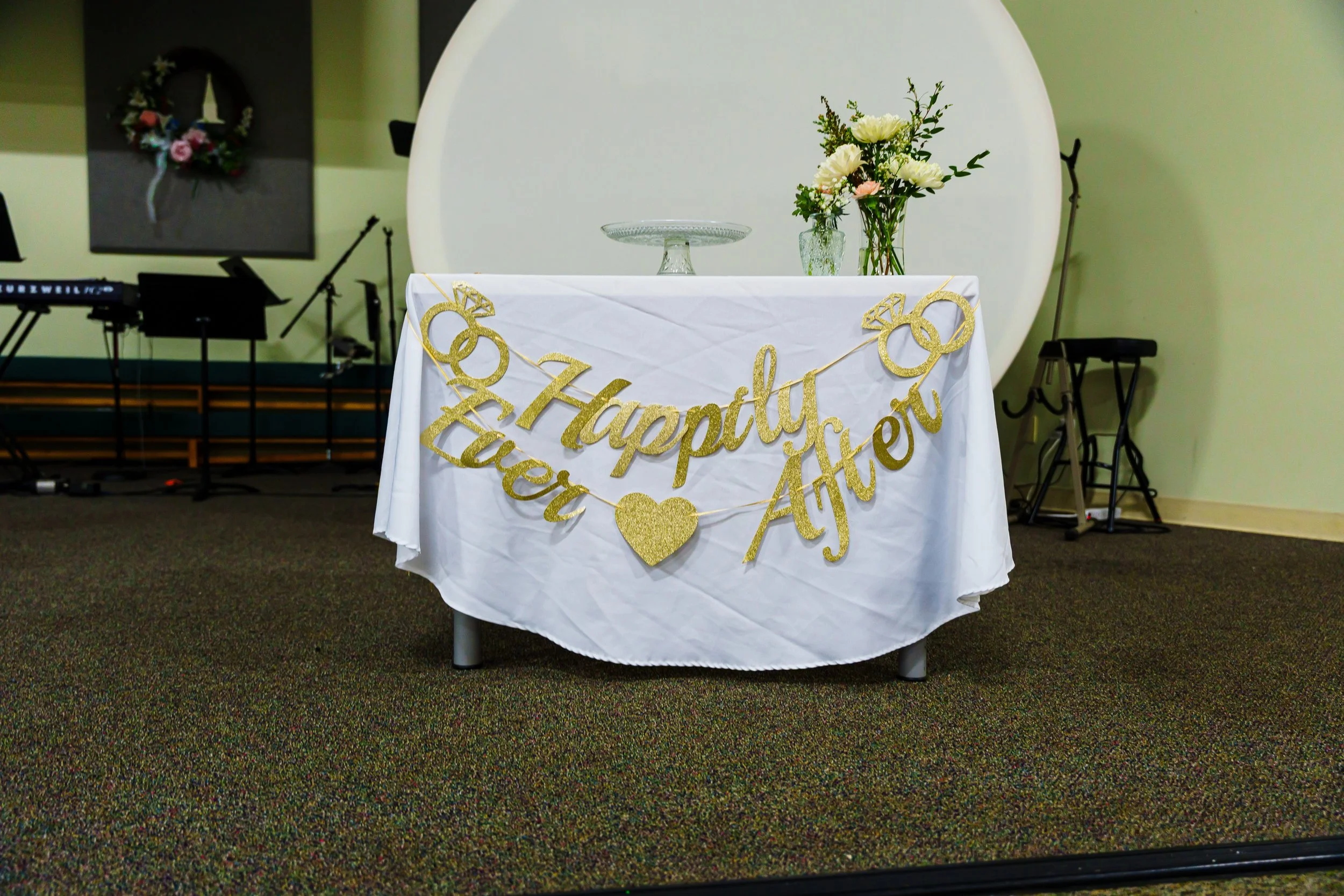 A table decorated for a wedding with a white tablecloth and a gold glitter banner reading "Happily Ever After". There are three vases with white and pink flowers on the table. In the background, there is a large white circular backdrop and a music ke