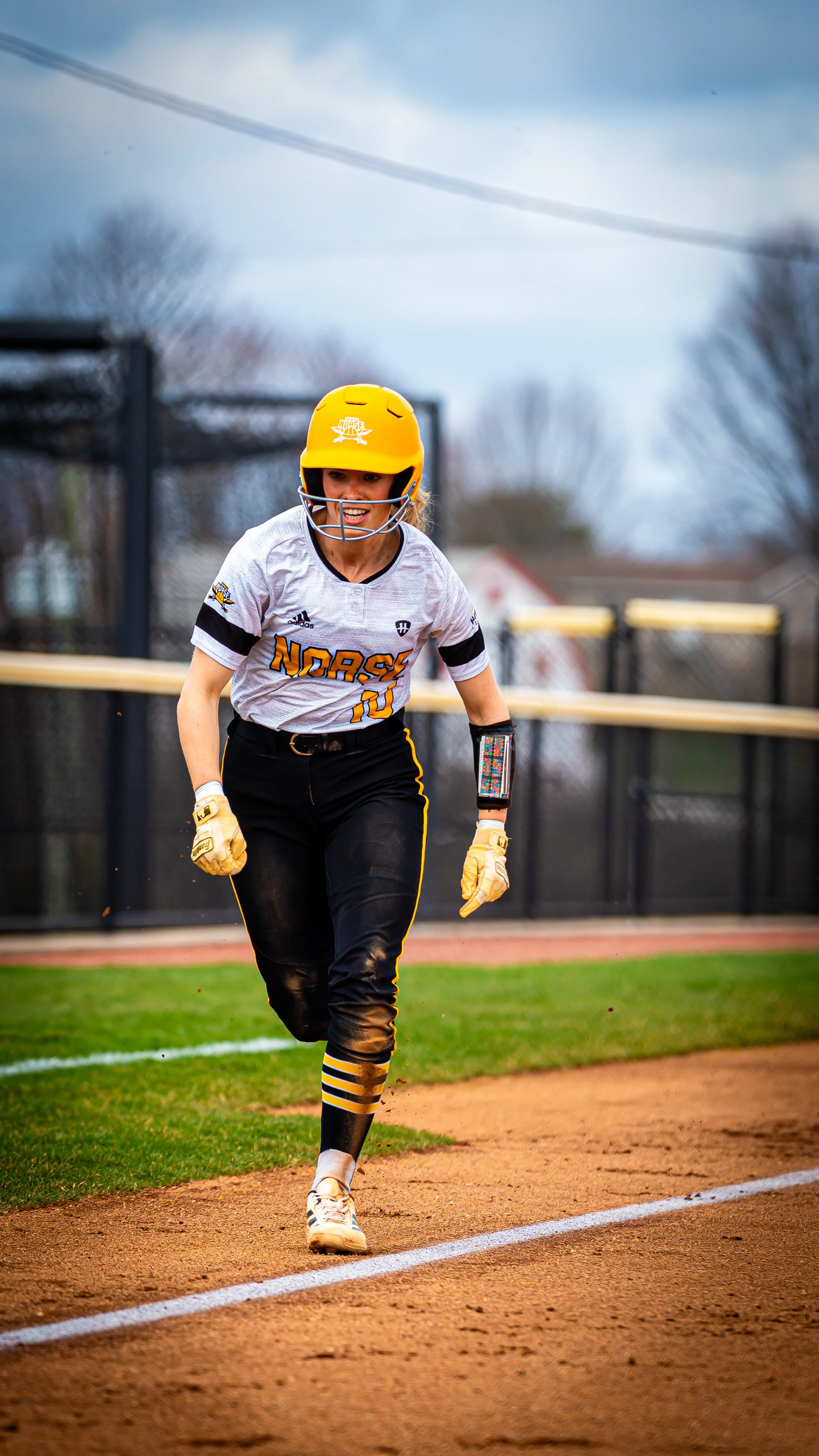 Female softball player running on the field during a game in the daytime, wearing a yellow helmet, white jersey with black and yellow accents, black pants, and striped socks.