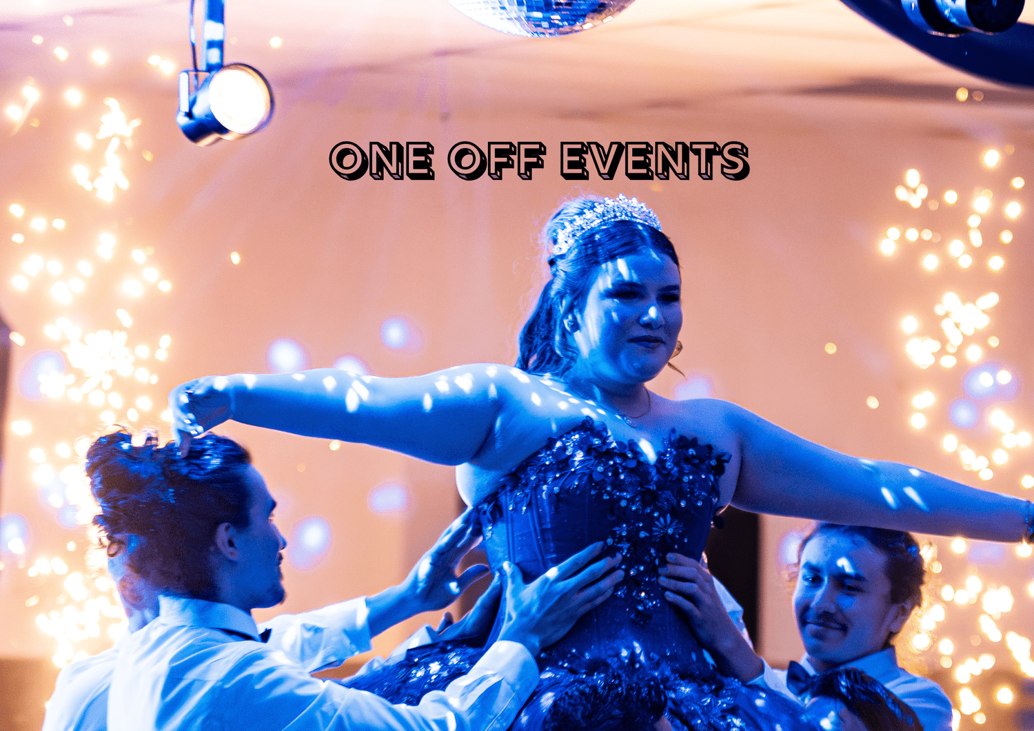 People dancing at a celebration, with a woman wearing a tiara and a sparkly dress being lifted by others, surrounded by colorful lights and sparklers.