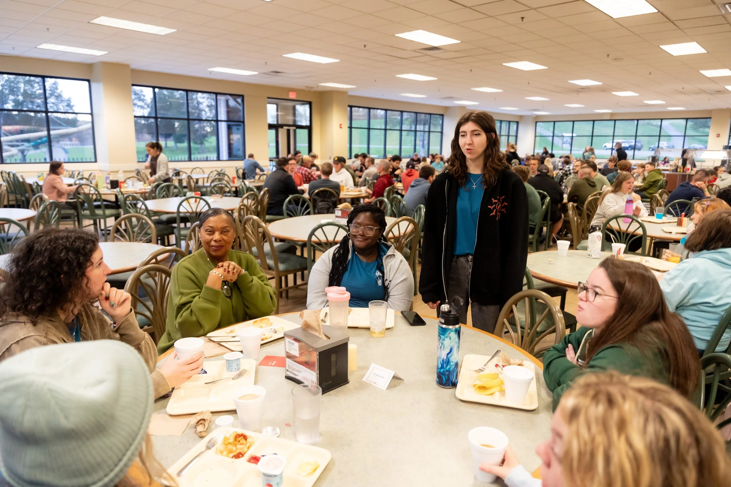 Group of people gathered in a large indoor dining area with large windows, sitting at round tables, eating and talking, with some standing and serving food.