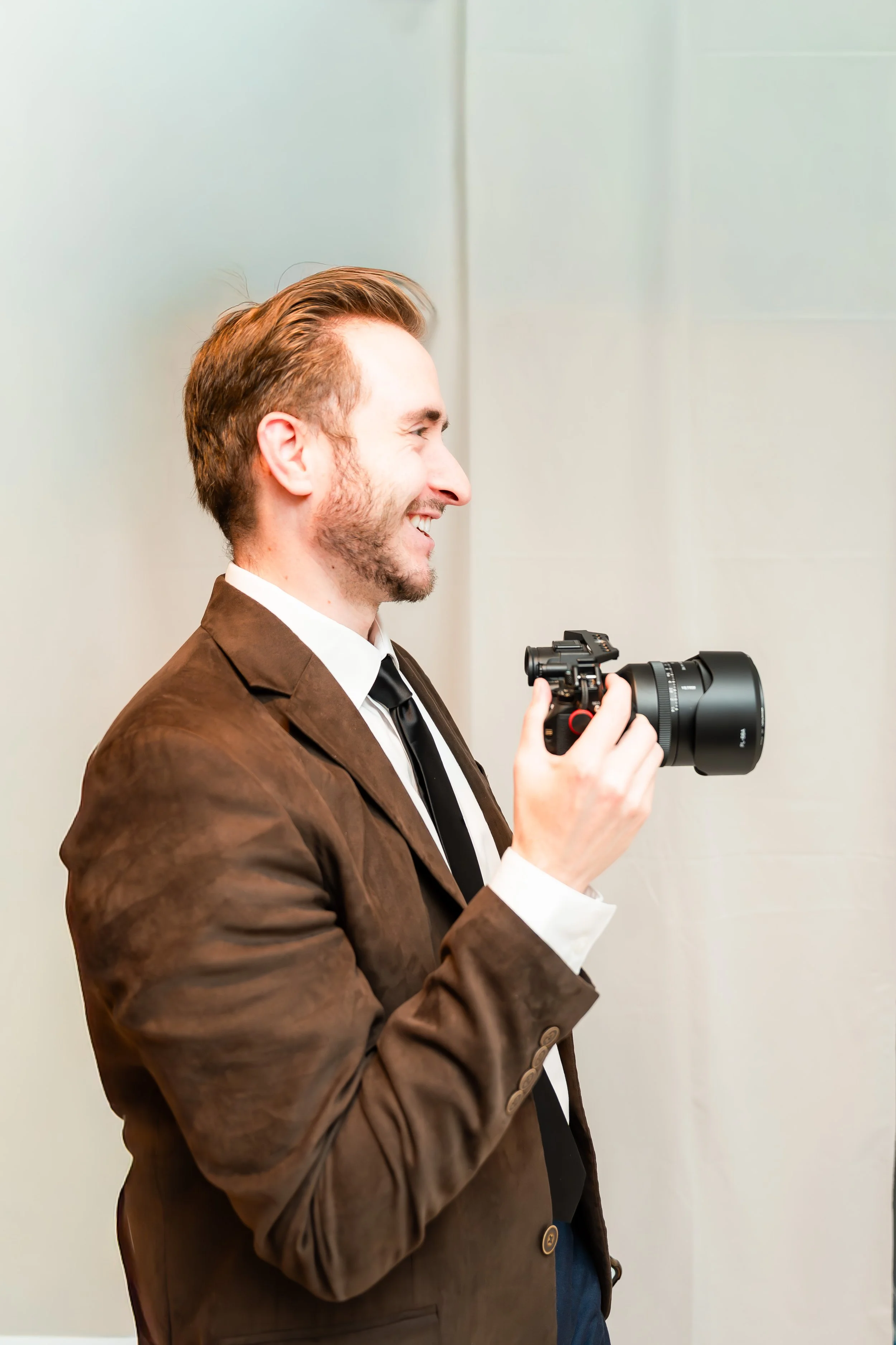 A man with reddish-brown hair and a beard, wearing a brown blazer, white shirt, and black tie, is smiling and holding a camera with a telephoto lens.