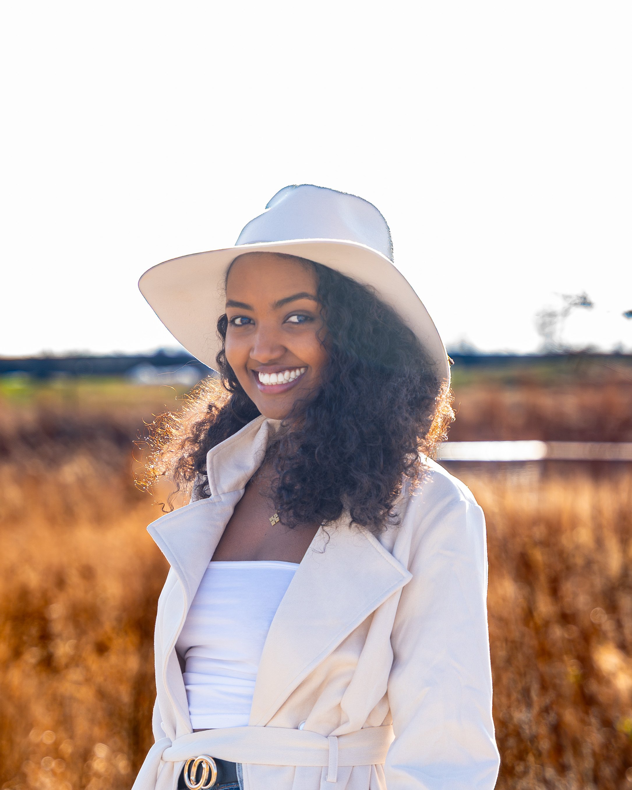 A woman smiling outdoors wearing a white wide-brimmed hat and a light-colored trench coat.