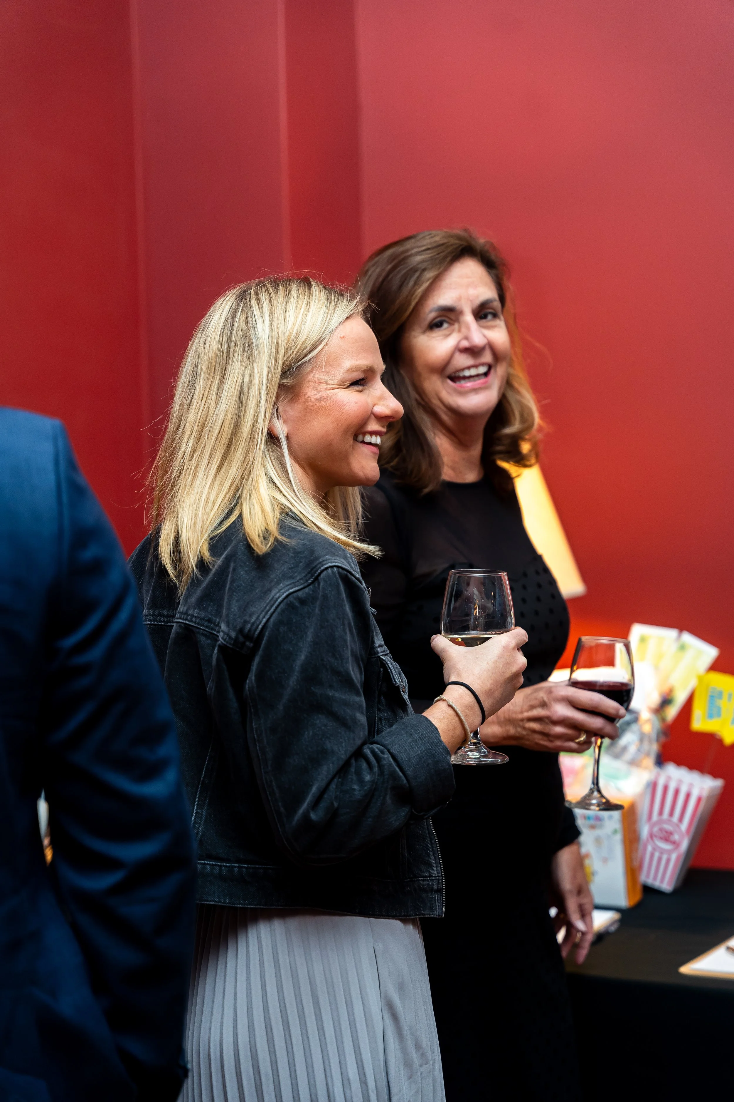 Two women smiling and holding glasses of red wine at a social gathering against a red wall decorated with colorful snacks and popcorn.