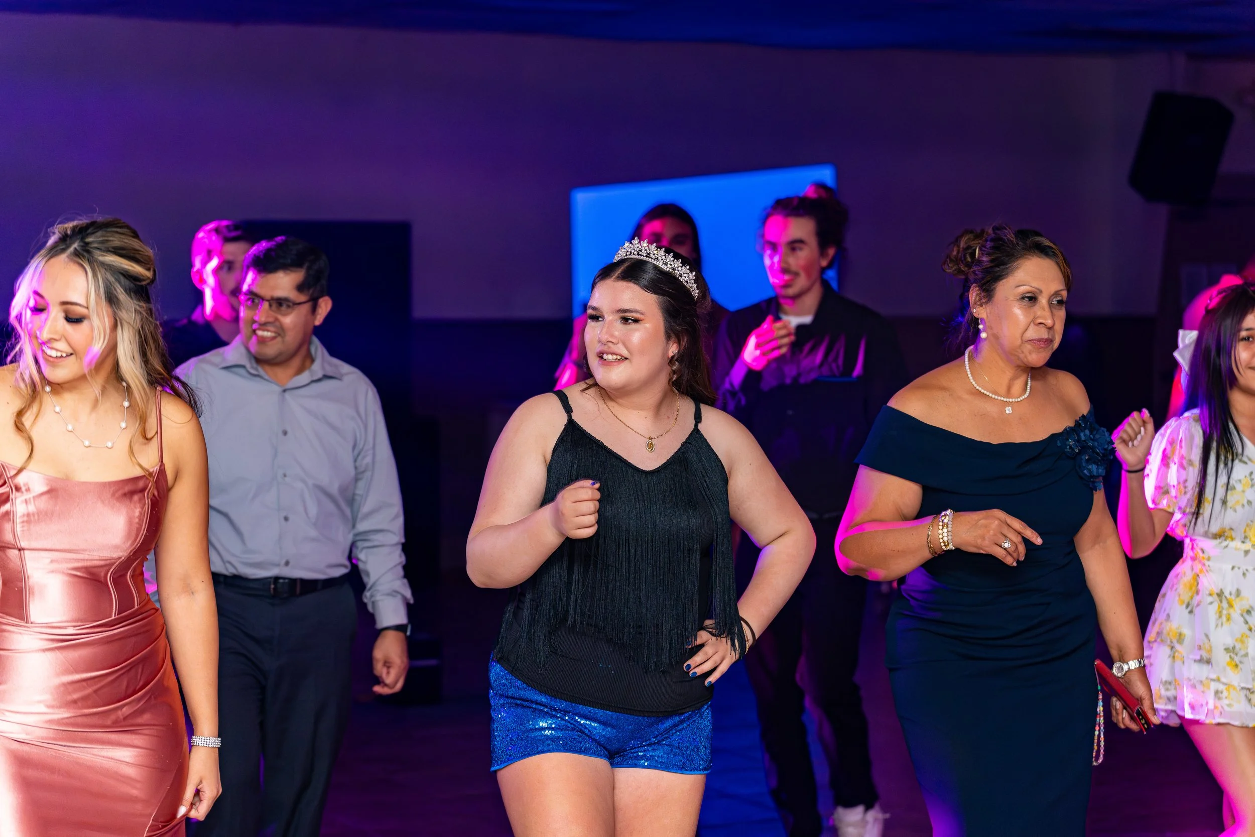People dancing at a celebration with purple and blue lighting, one woman wearing a tiara in the center.