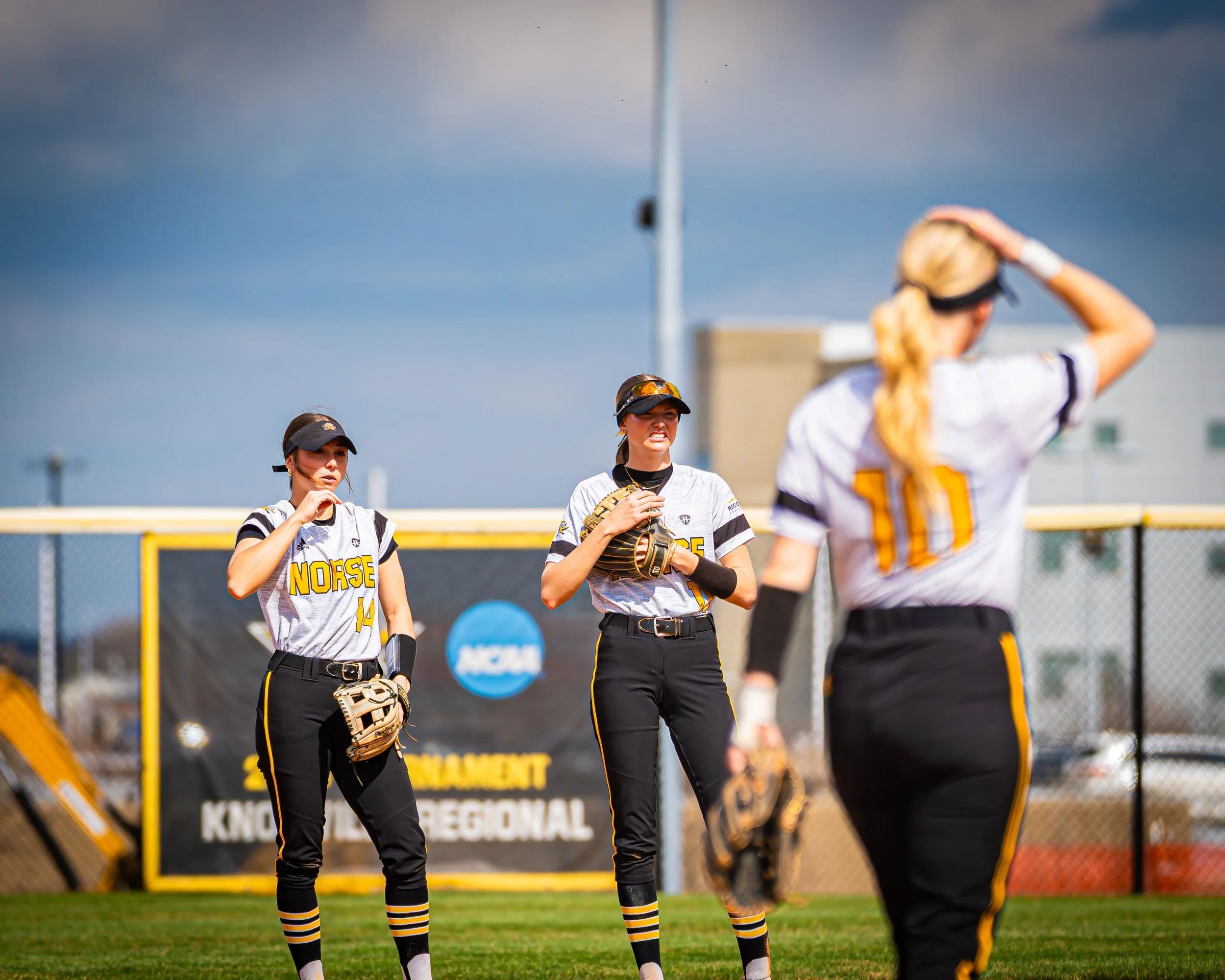 Girls softball team in uniforms and caps standing on a field before a game, with a yellow fence and a sign in the background.
