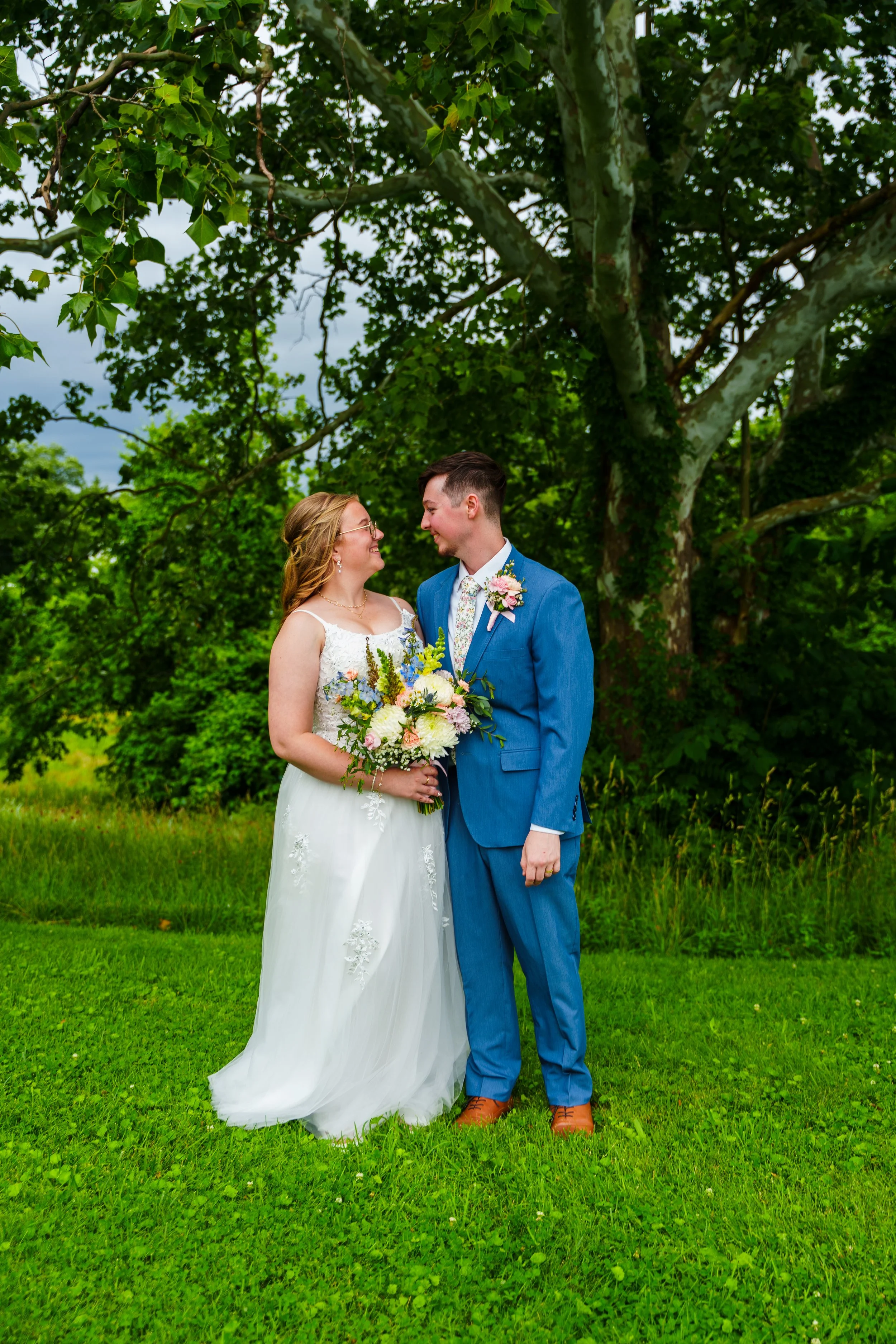 A bride and groom standing close together outdoors on green grass, smiling and looking at each other, with trees in the background.