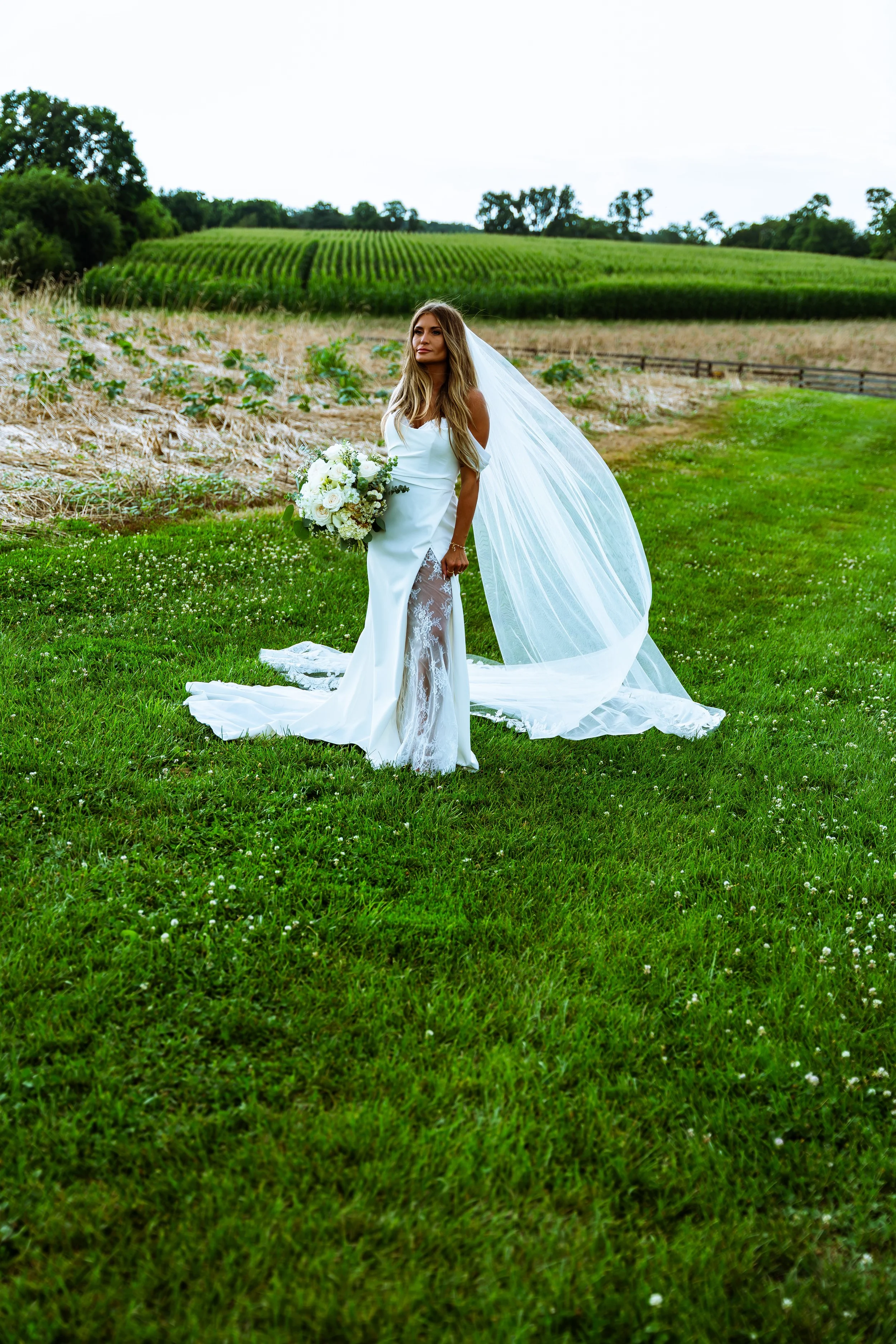 A woman in a white wedding dress with a veil holding a bouquet of white flowers standing on lush green grass in a rural field with rows of crops and trees in the background.