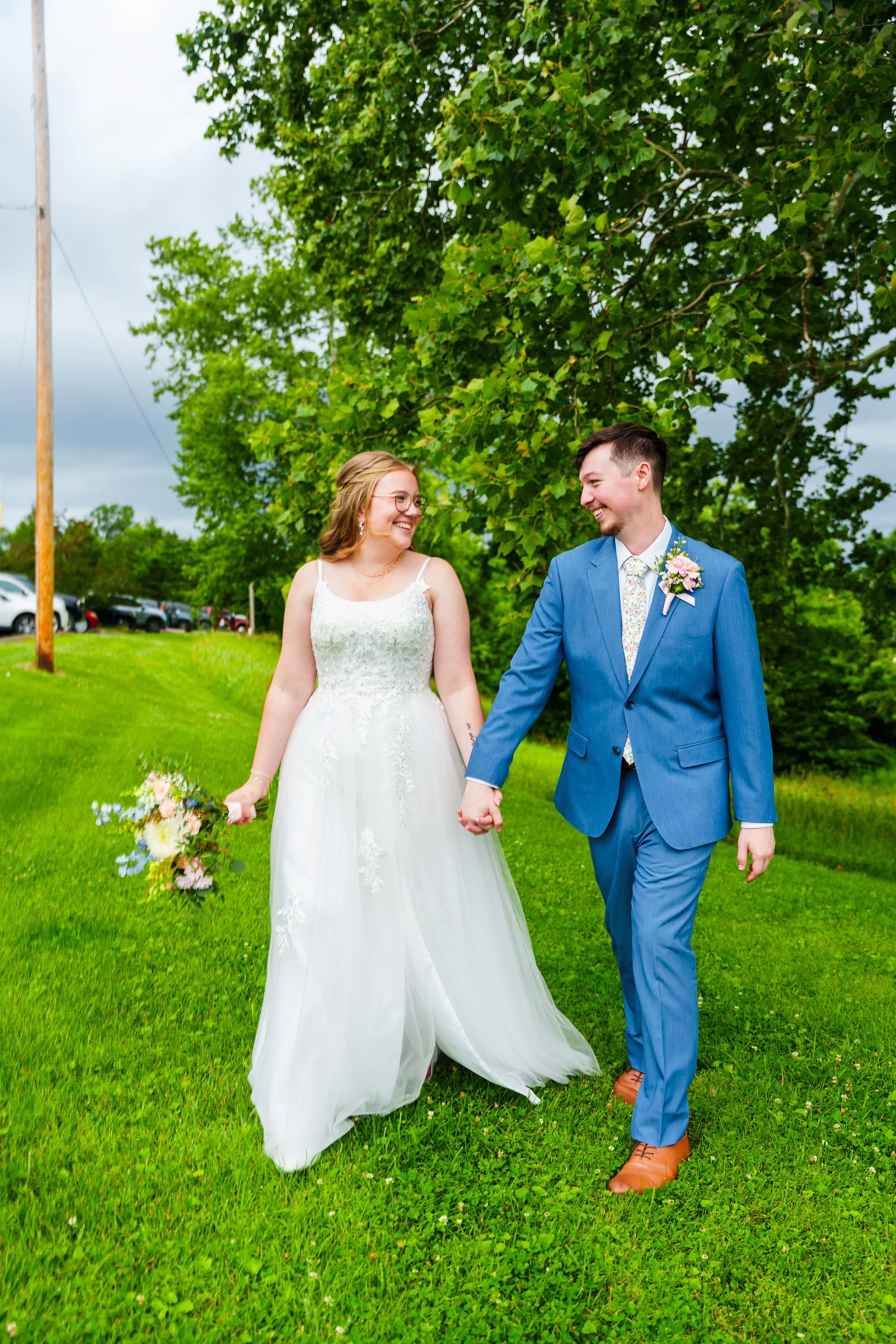 A bride and groom holding hands and smiling at each other outdoors on green grass with trees and parked cars in the background.