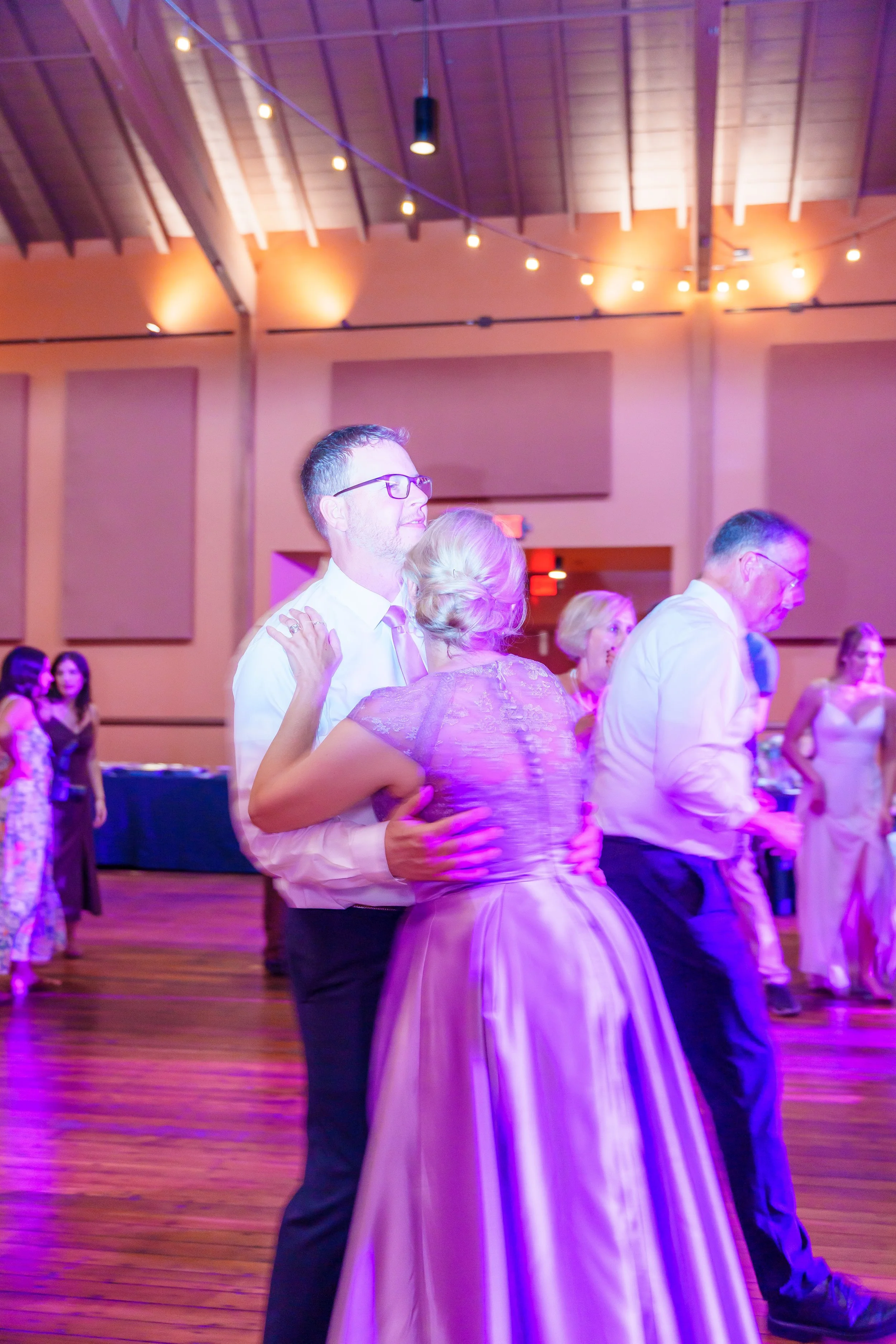 People dancing at a social event in a large wooden hall with string lights overhead