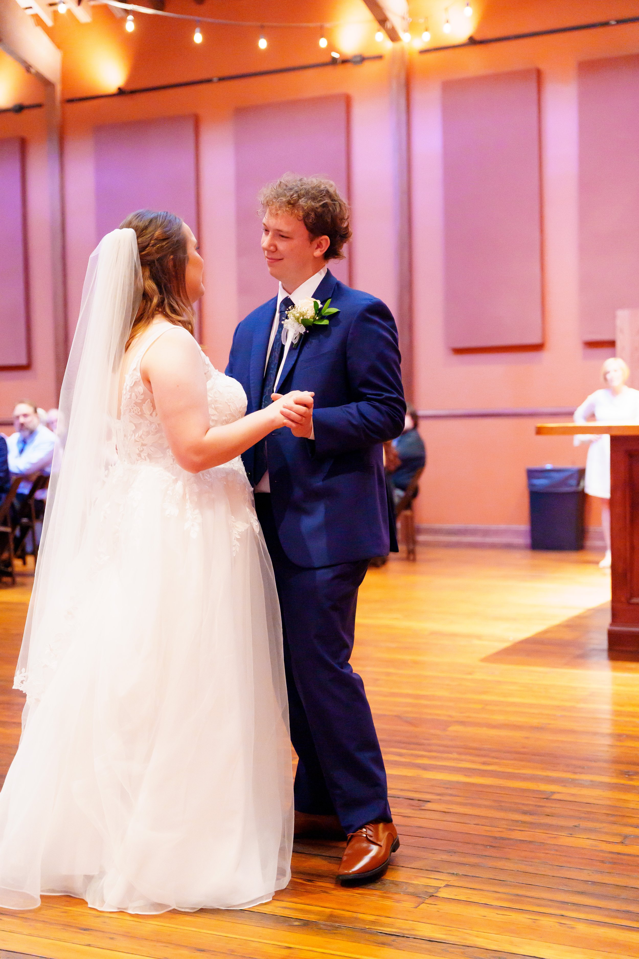 A couple dancing at their wedding in a warmly lit indoor venue. The bride is in a white lace wedding gown with a veil, and the groom is wearing a navy blue suit with a white shirt and tie. They are holding hands and gazing at each other. The backgrou