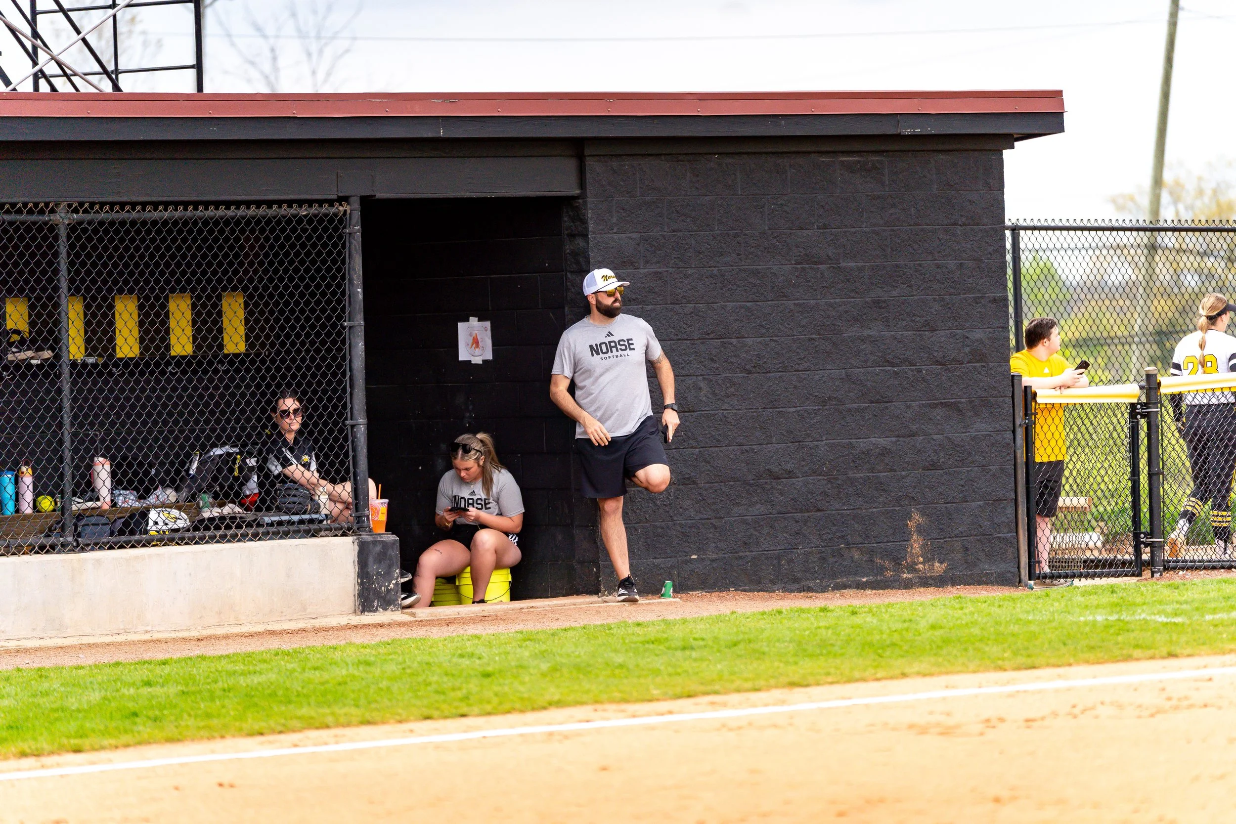 People sitting and standing near a dugout at a softball game, with some wearing yellow jerseys and one person wearing a gray shirt that says 'NOSE SOFTBALL.'