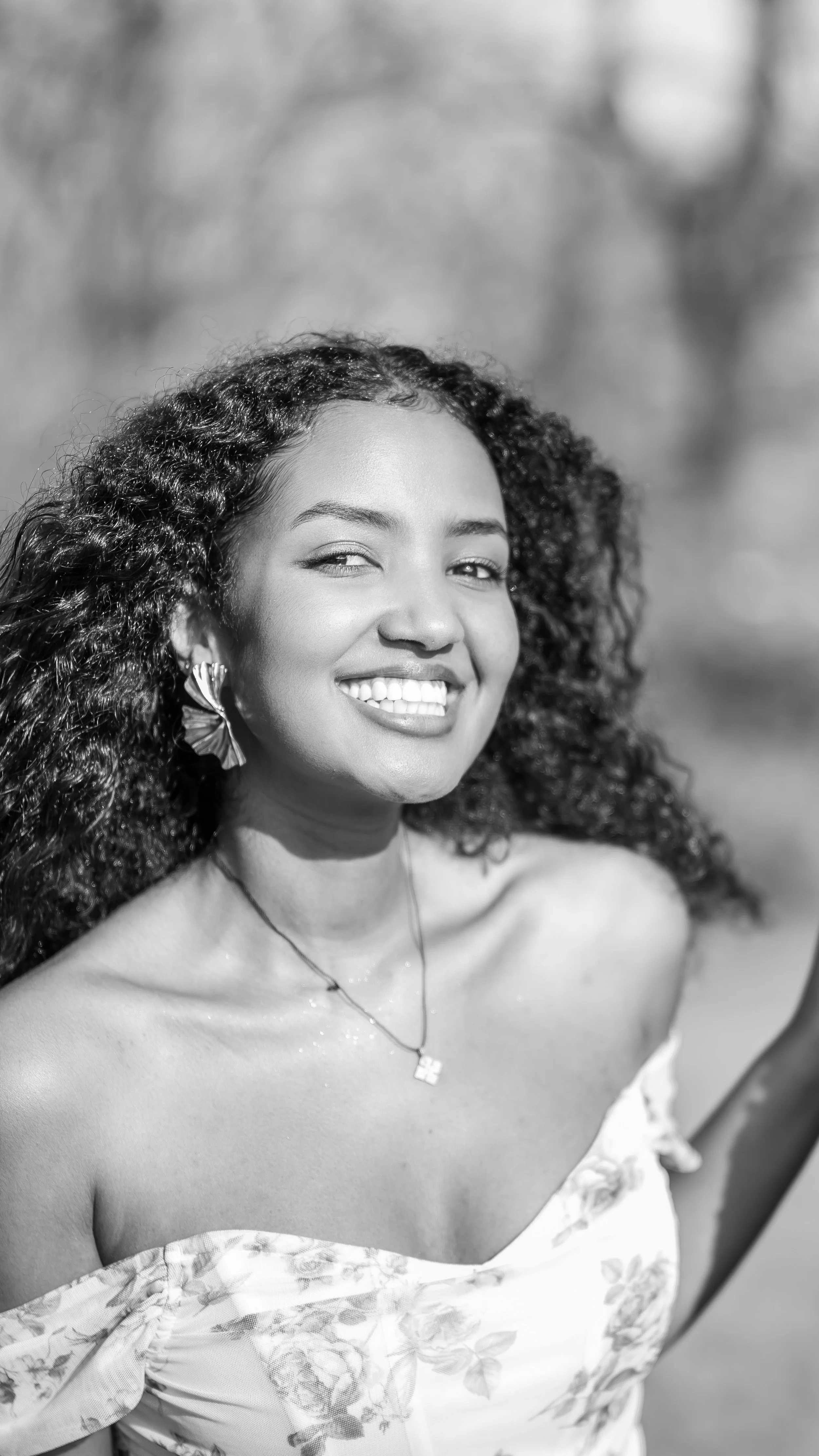 Black and white photo of a smiling woman with curly hair, sitting outdoors, wearing an off-the-shoulder floral top and jewelry.