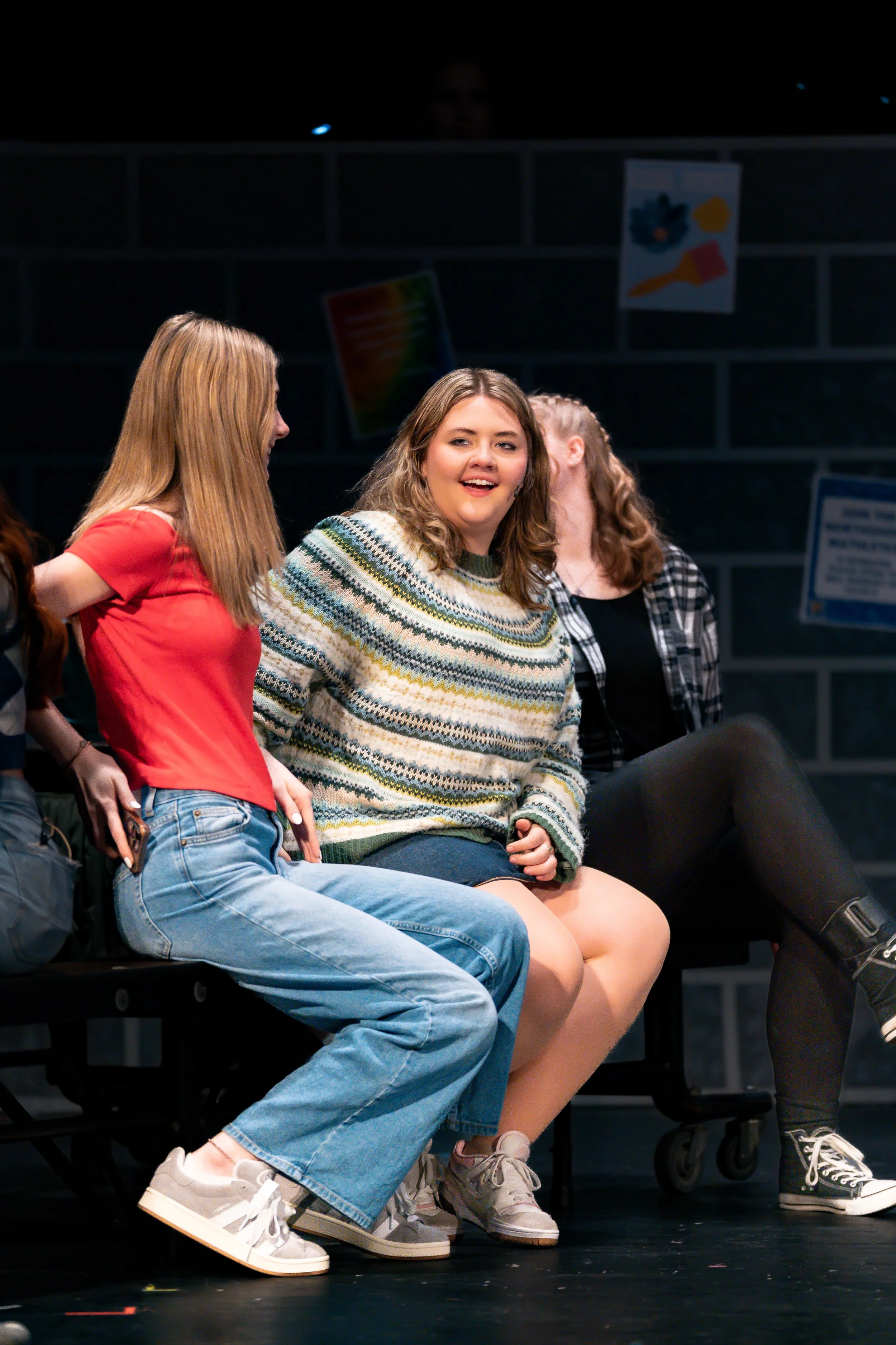 Three young women sitting on chairs in a conference or presentation setting, engaging in conversation and smiling. The background shows a black brick wall with some posters and signs.