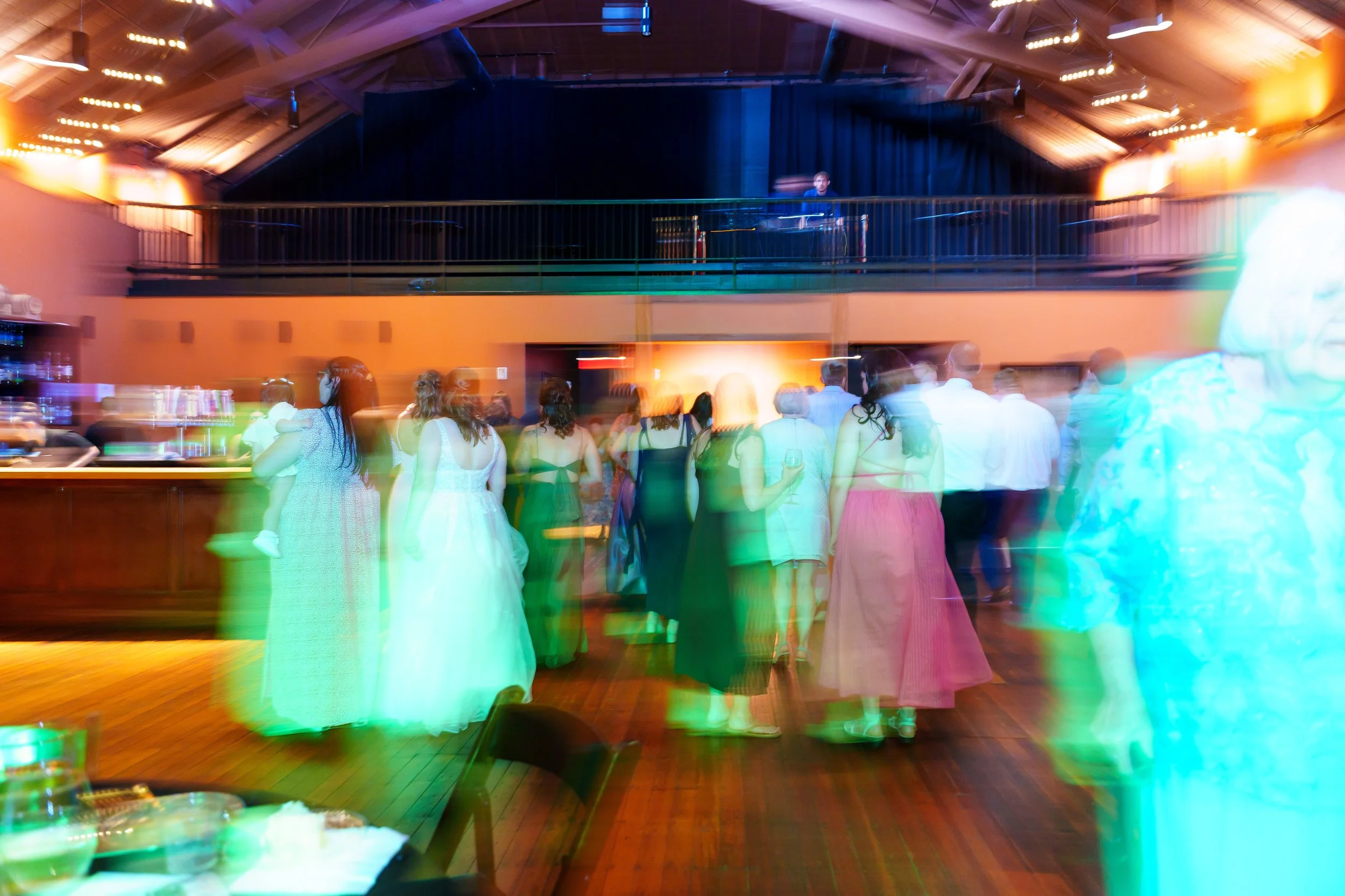 A lively party scene with people dancing in formal attire inside a decorated venue, featuring a DJ or musician on a raised platform in the background, and colorful lighting effects creating a dynamic atmosphere.