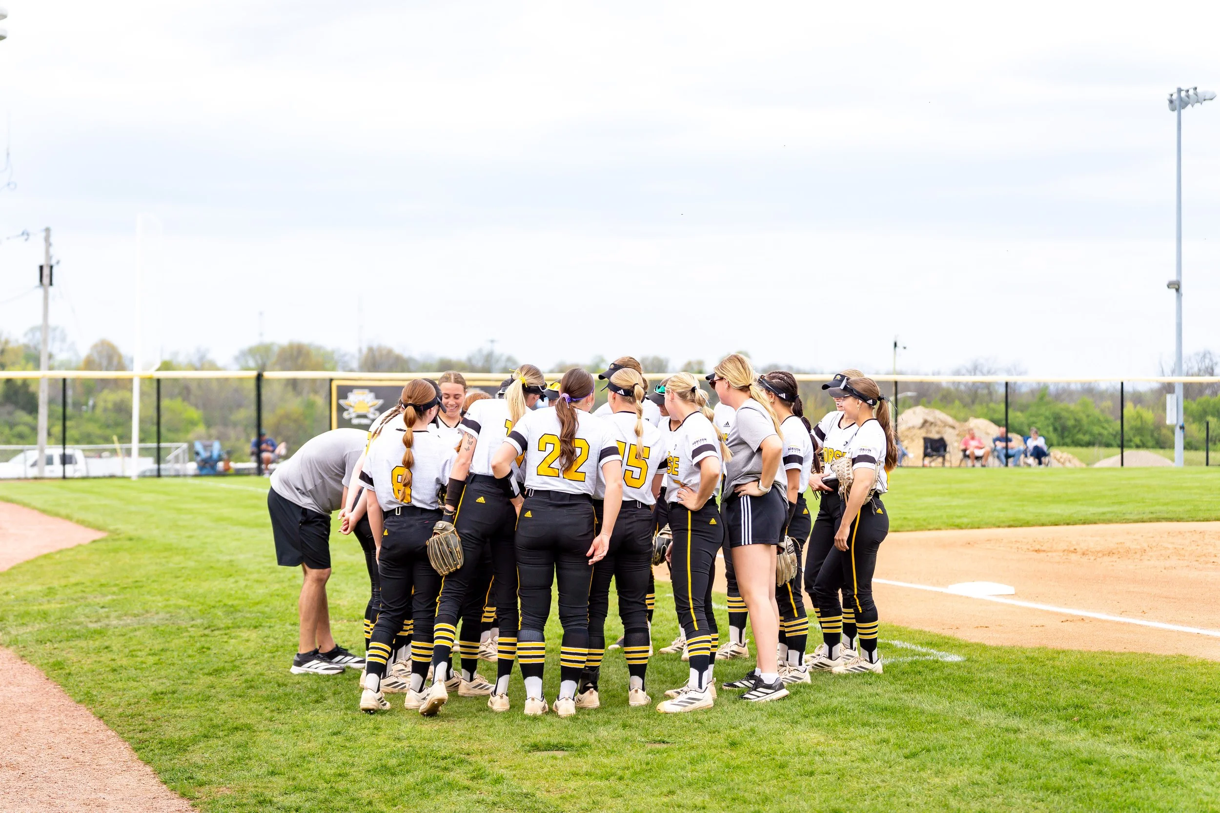 A group of female softball players in white and black uniforms gathered in a huddle on a baseball field.