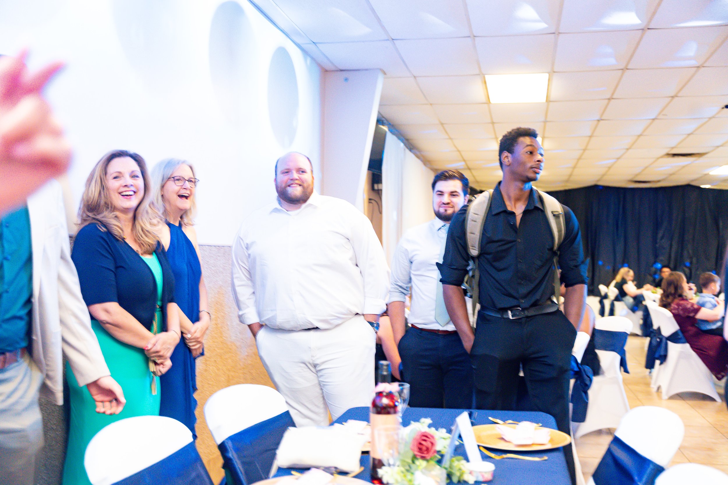 Group of people standing and smiling at a formal event or gathering in a banquet hall with decorated tables and chairs.