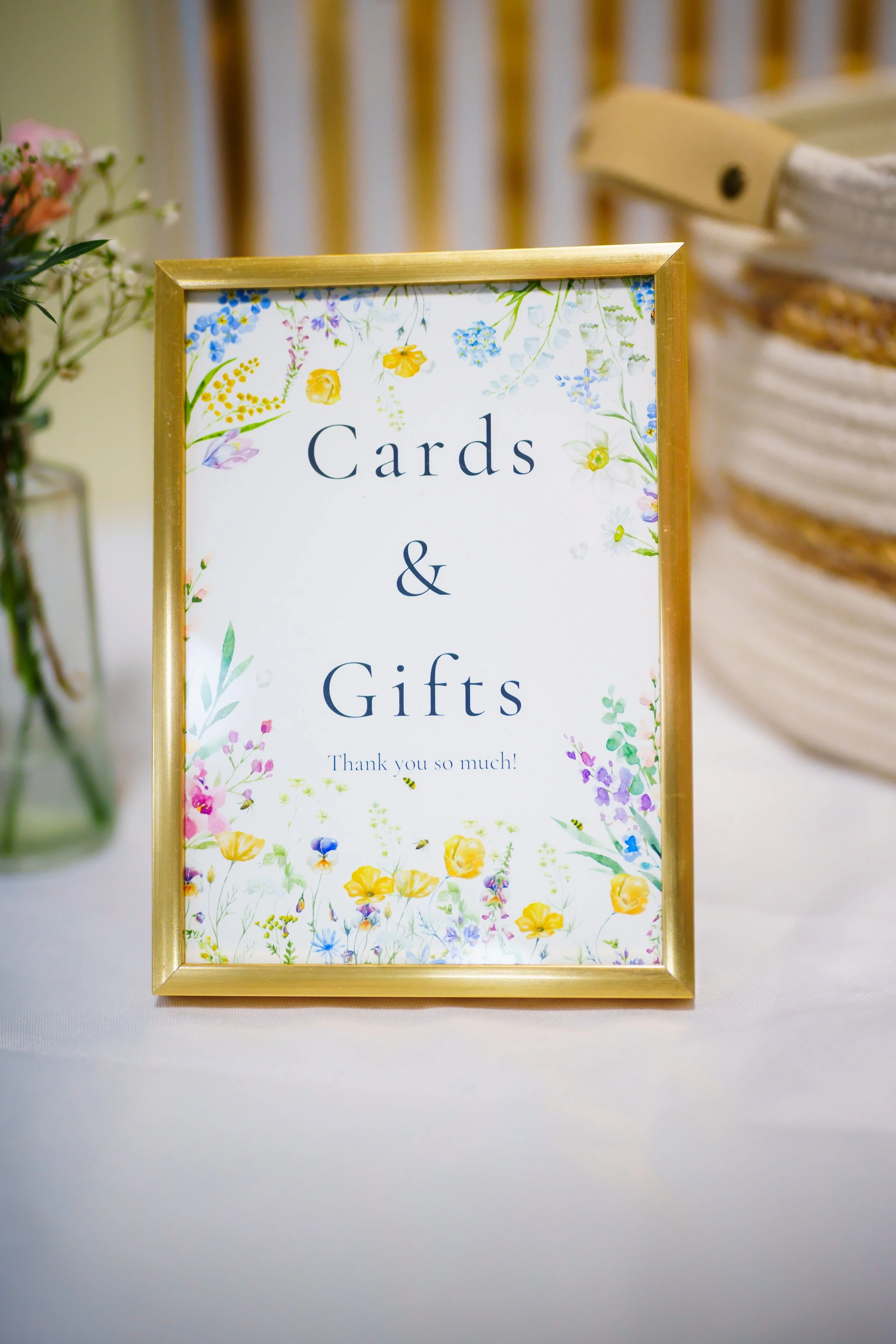 A framed sign with floral design that says 'Cards & Gifts' with a thank you message, positioned on a white table with a blurred basket in the background.