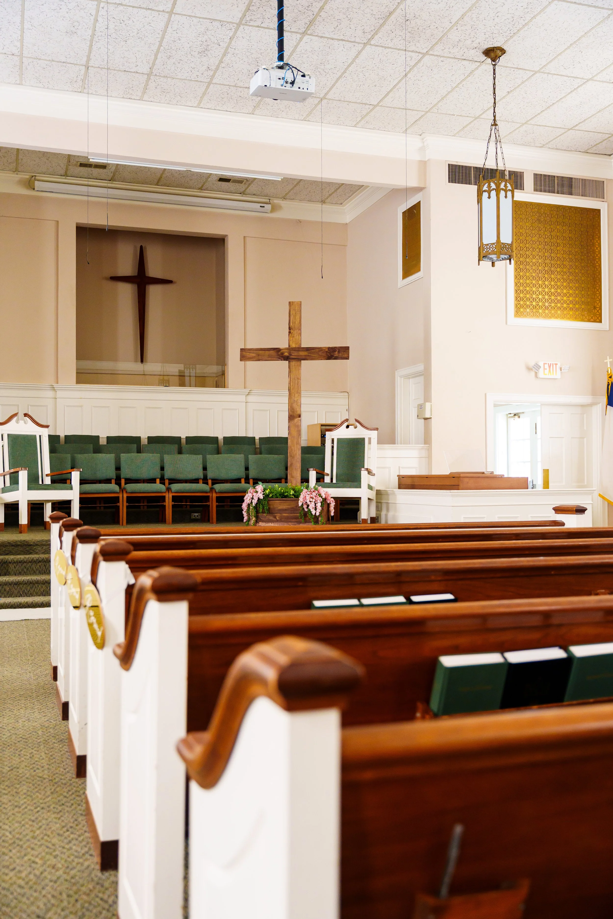 Interior of a church sanctuary with wooden pews, a large wooden cross at the front, green upholstered chairs, floral arrangement, and a hanging lantern light fixtures.