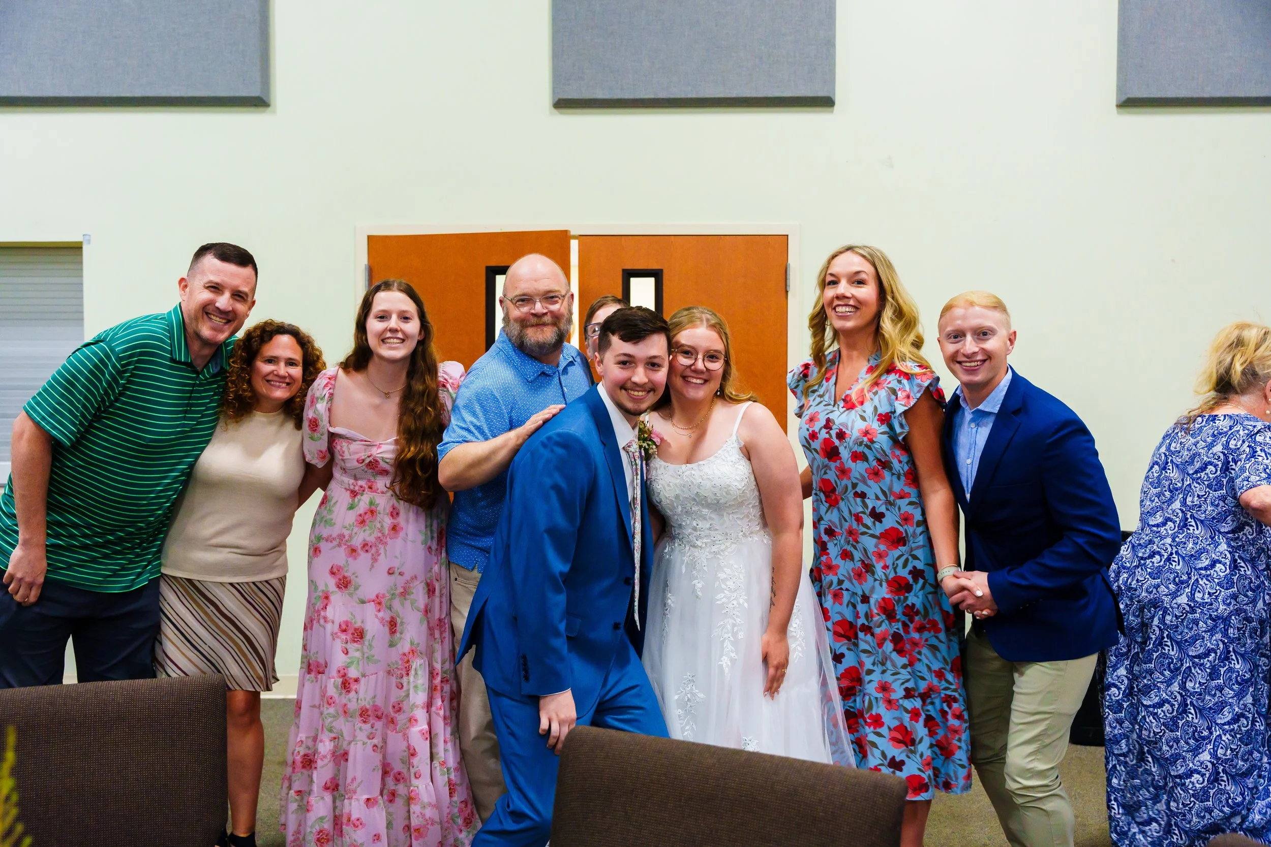 A group of ten people posing indoors near a closed door, dressed in formal and semi-formal attire, smiling for a photo at a celebration or event.