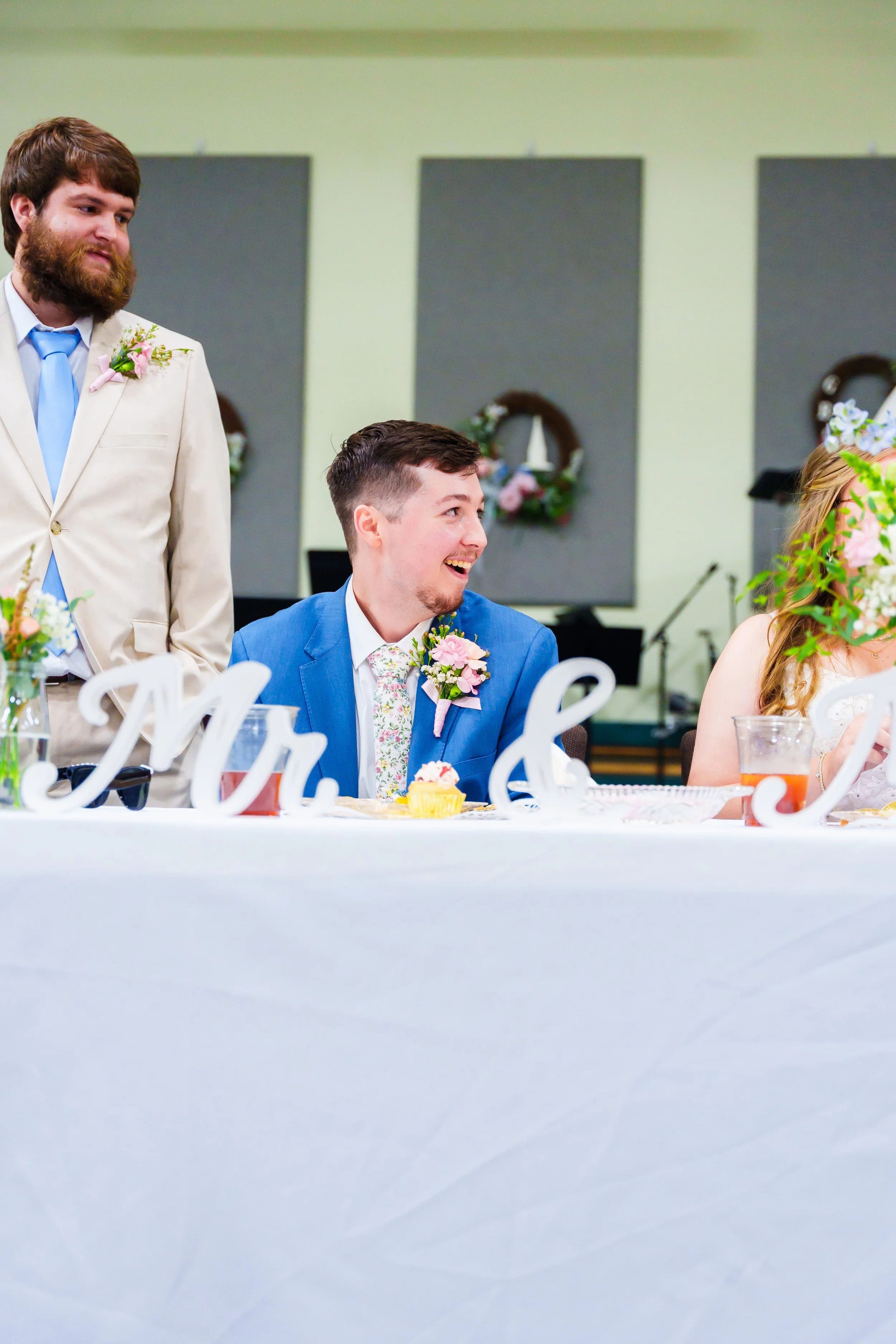 People at a wedding reception, seated at a decorated table with floral arrangements and fondant words, with a man in a blue suit smiling.
