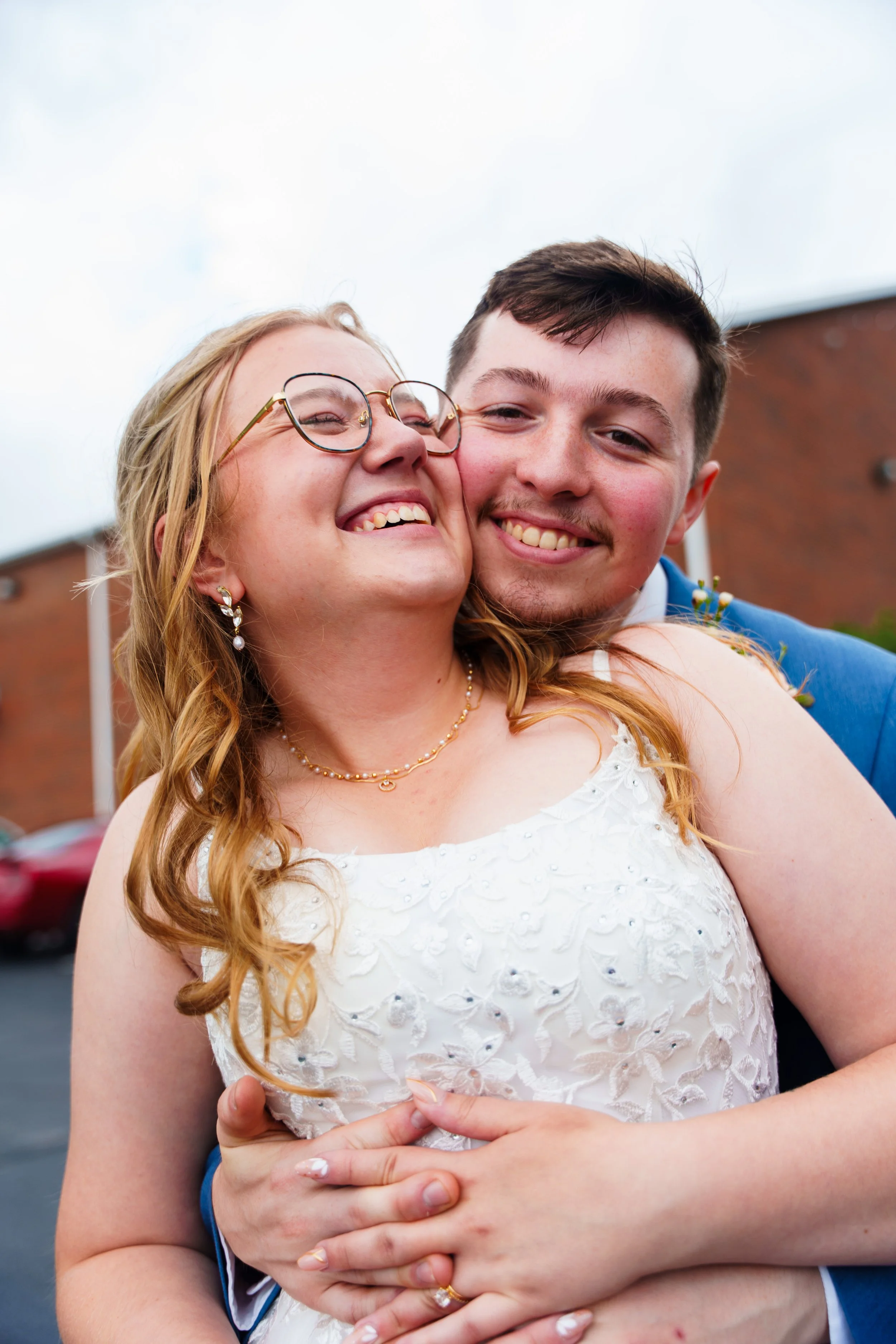 A happy couple embraces, with the woman smiling widely and the man smiling softly. The woman is wearing glasses, jewelry, a white dress with lace details, and has long, curly hair. The man has short, dark hair and is dressed in a blue jacket. They ar