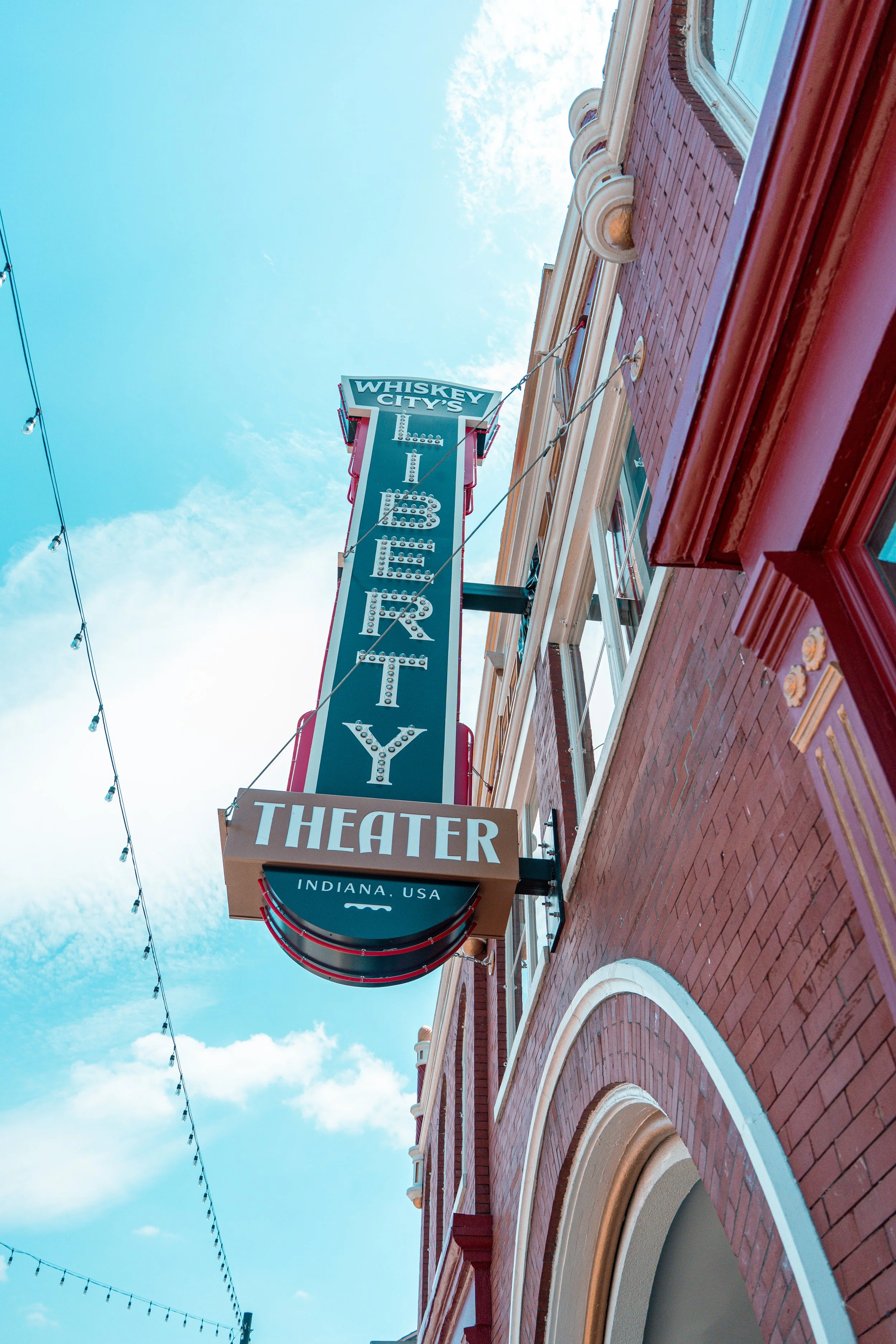 A vertical sign for Whiskey City Theater in Indiana, USA, hanging on a red brick building with white decorative borders and windows, with string lights against a partly cloudy blue sky.