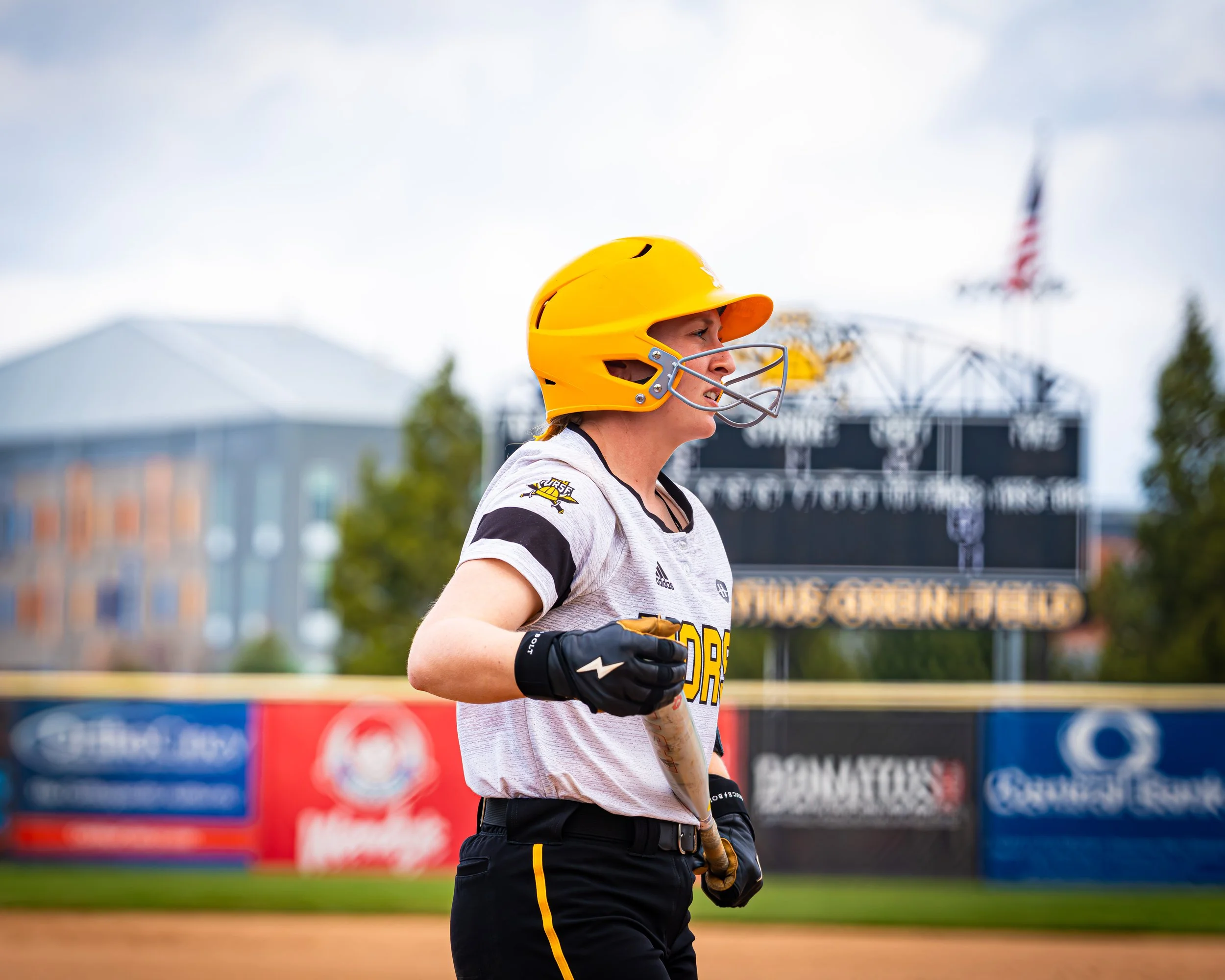 Young woman in baseball uniform and yellow helmet standing on baseball field.
