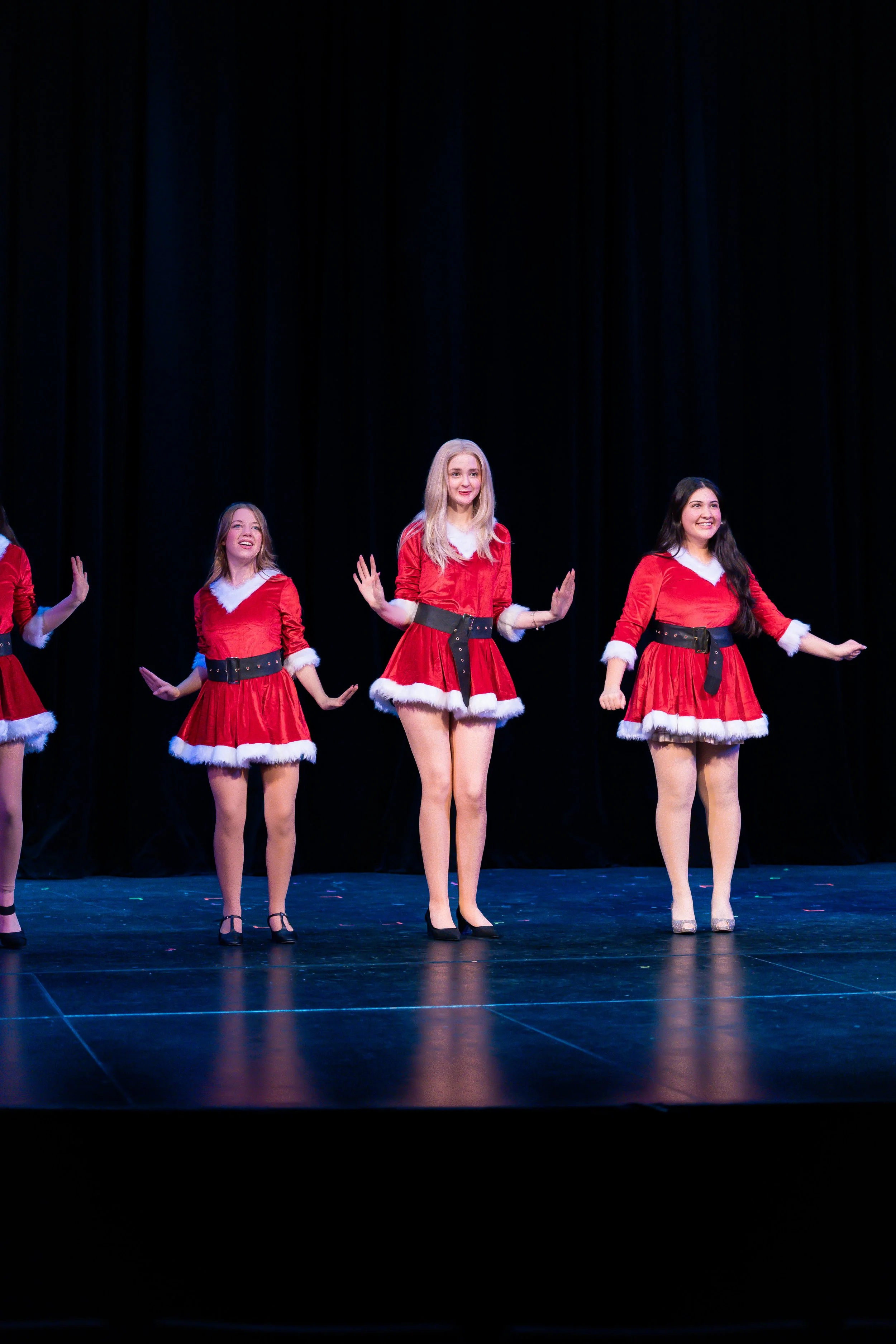 Four young women dressed in Christmas-themed red dresses with white fur trim, black belts, and heels are performing a dance routine on stage with a black curtain backdrop.