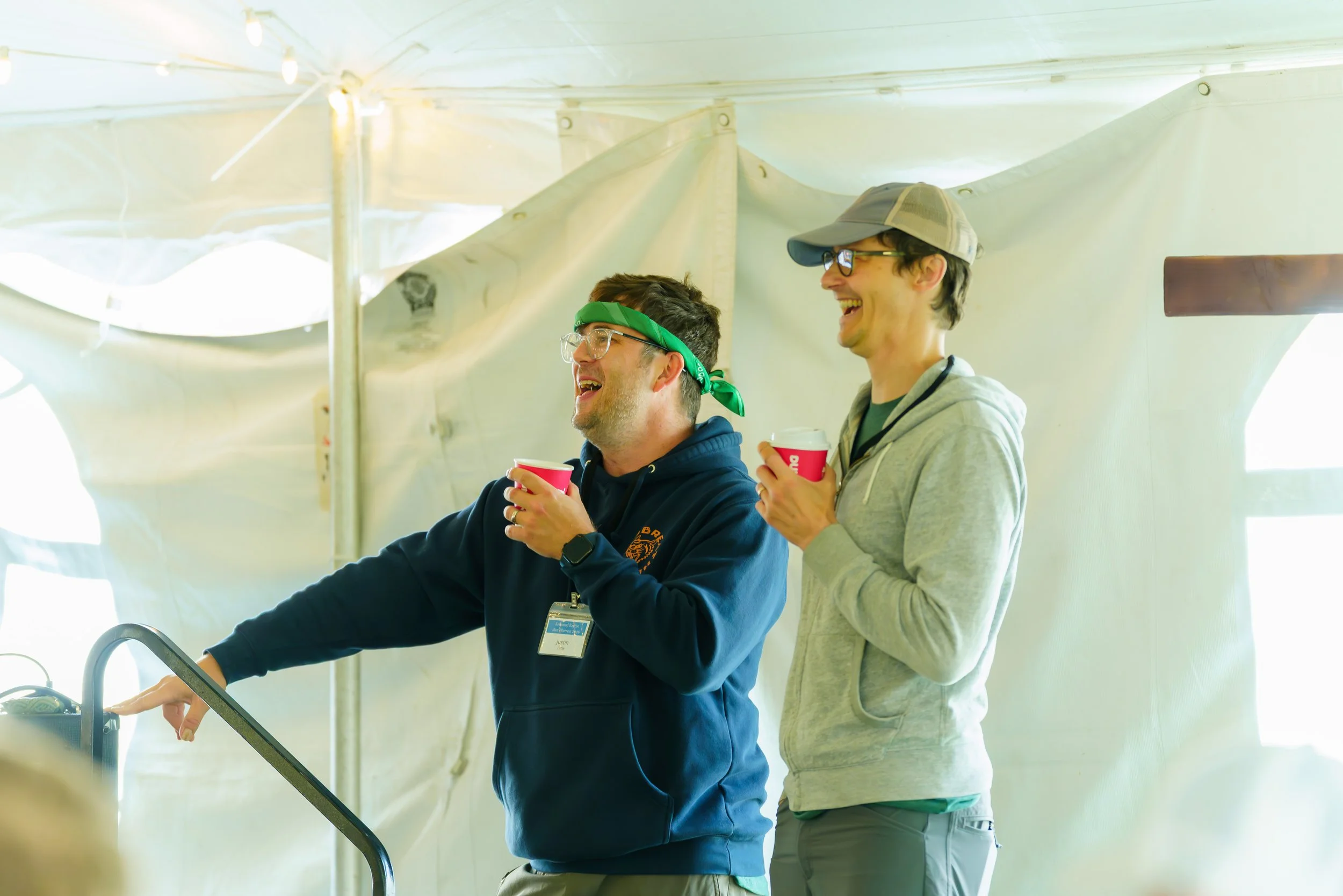 Two young men smiling, holding red cups, standing inside a tent or canopy. One is wearing glasses, a green bandana, and a dark hoodie with a name tag, while the other is wearing a cap, glasses, and a gray hoodie.