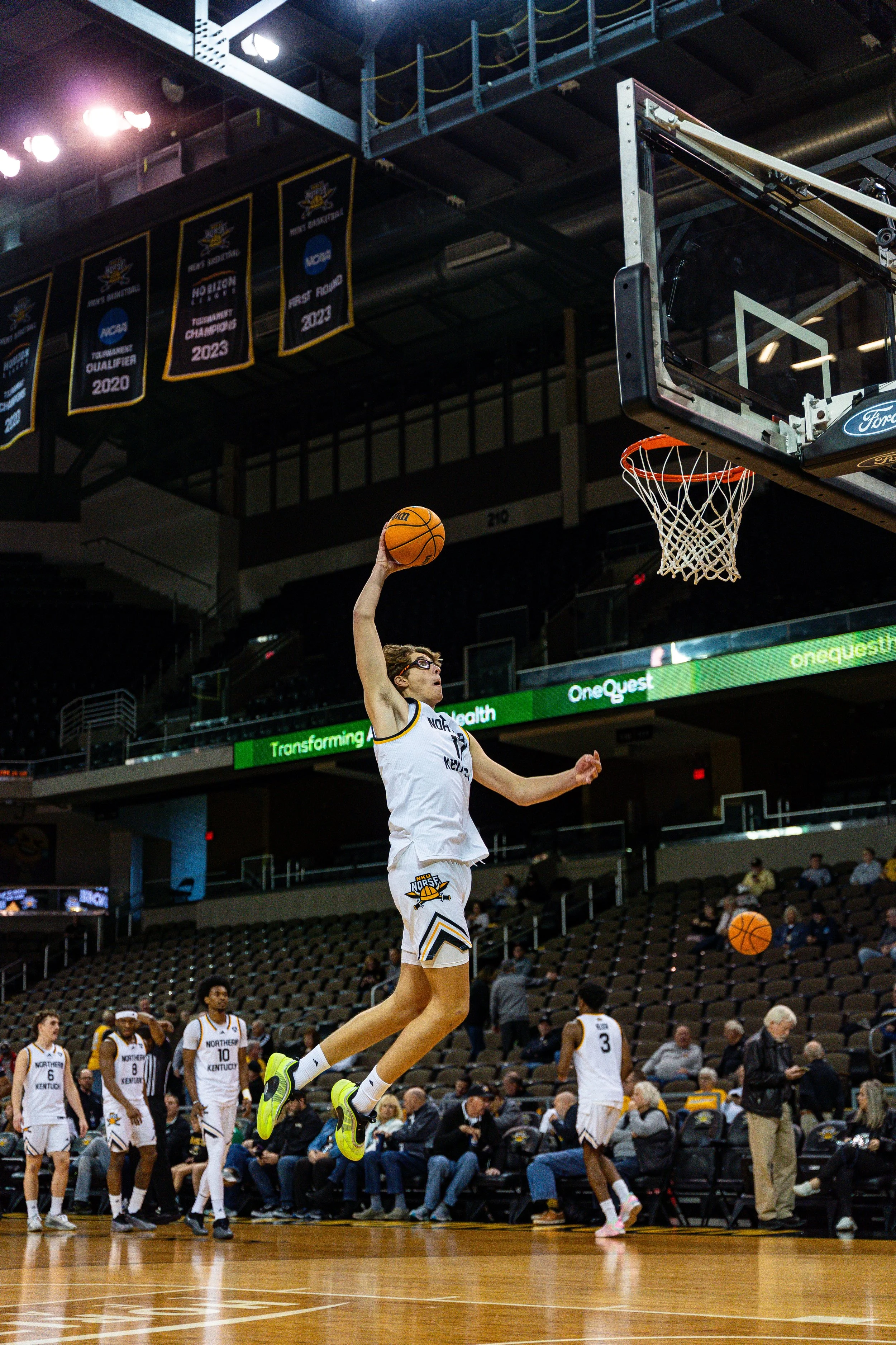 Basketball player in jersey number 12 jumping to score a basket during a game inside an arena, with teammates and spectators in the background.