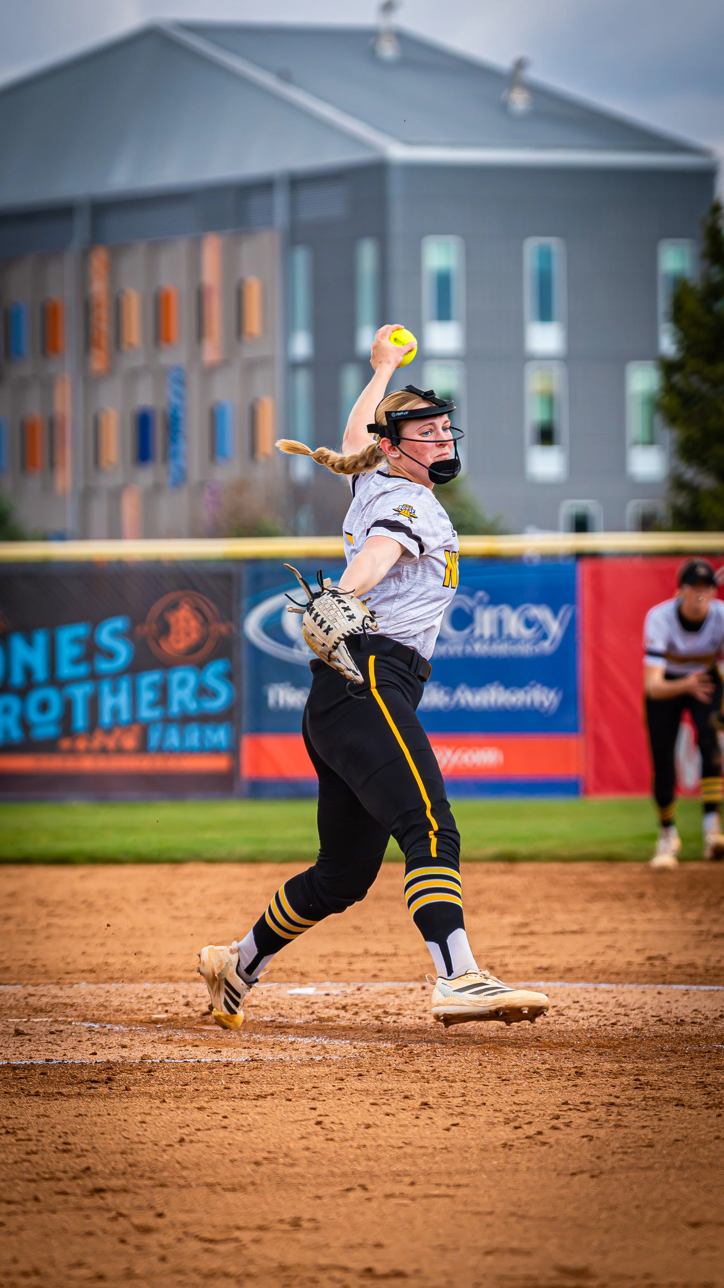 A female softball player in a gray uniform with black and yellow accents is preparing to throw a yellow softball on the field. She is wearing a protective face mask and glasses, with her hair in a braid. There is a building and a fence with advertise