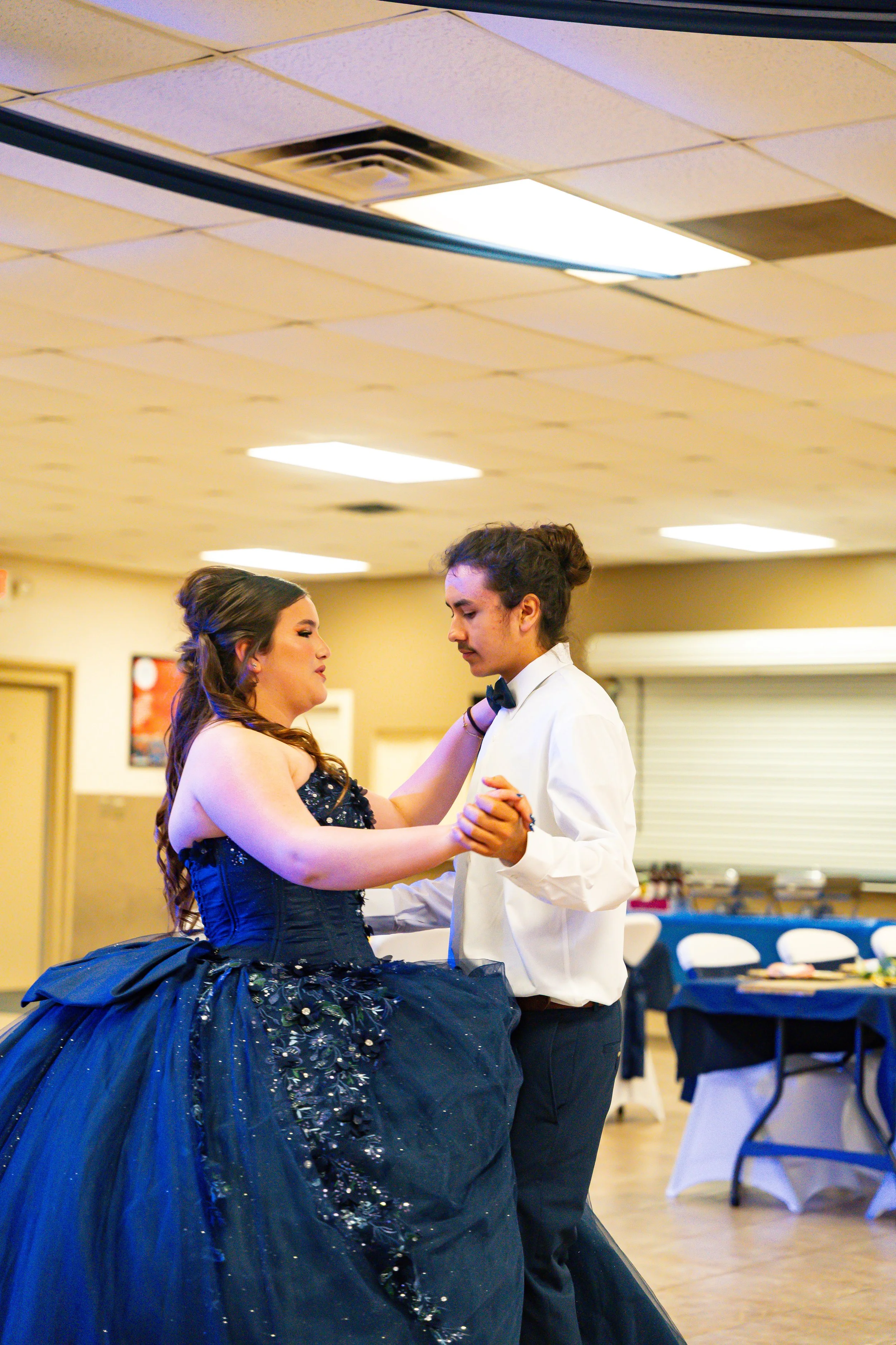 A young woman in a dark, shiny, strapless gown with floral embellishments dances with a young man in a white shirt and black bow tie at an indoor event. The woman has long, wavy hair, and they are holding hands while looking at each other.