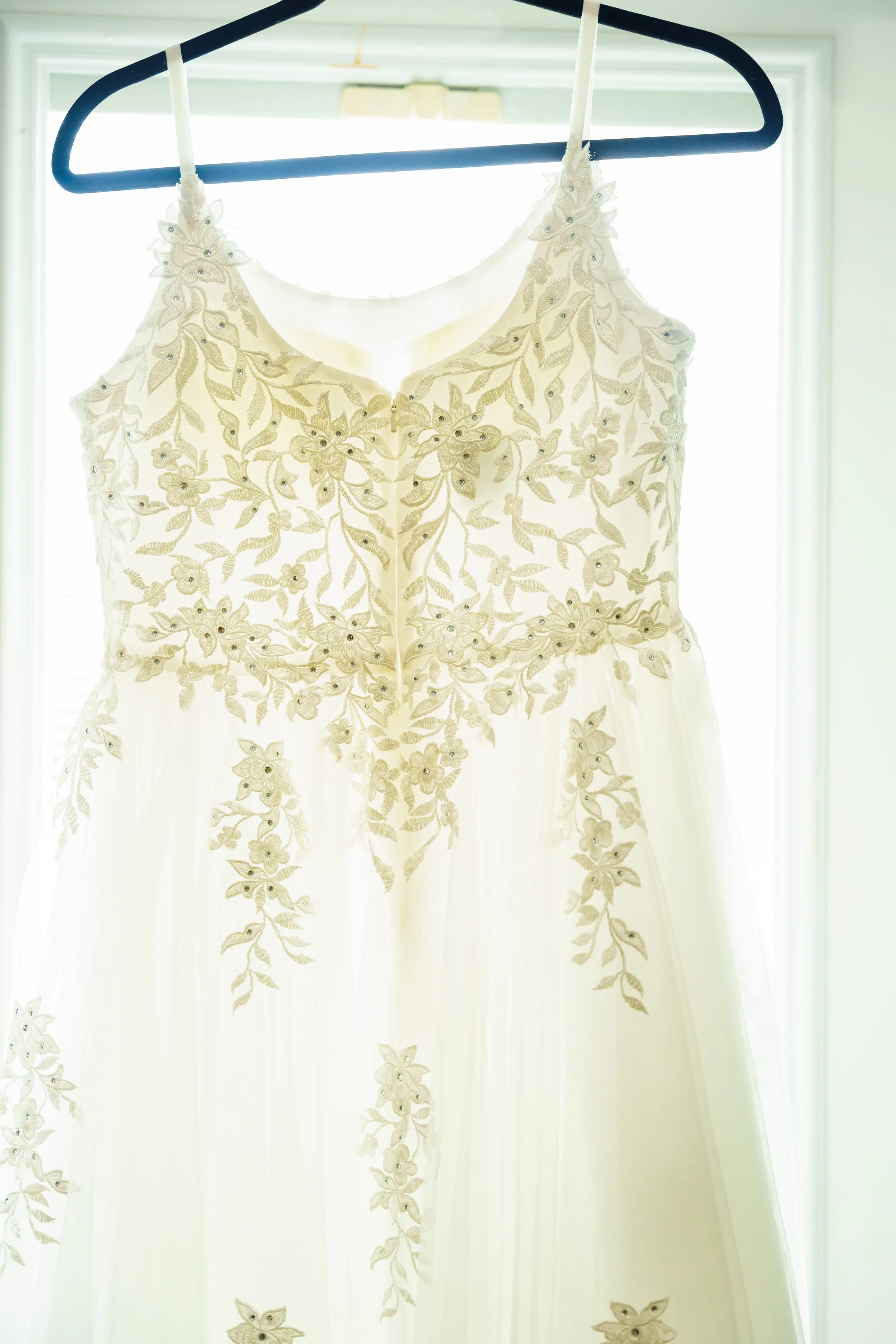 A wedding dress with intricate floral embroidery and bead details hanging on a black hanger in front of a window with bright light shining through.