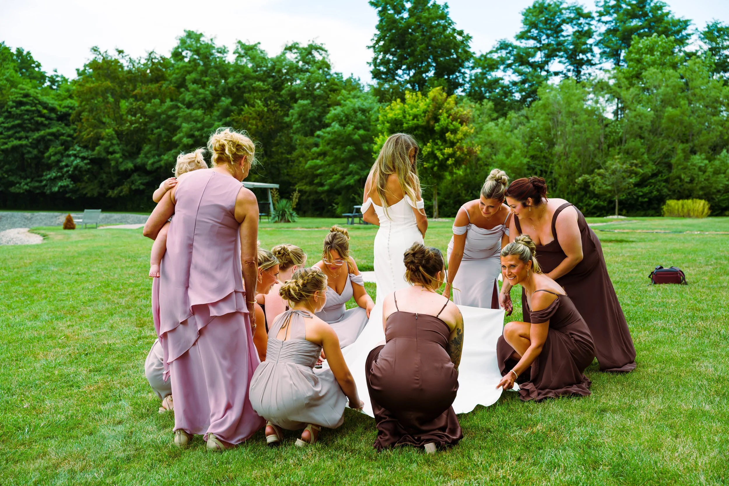 Group of women and girls dressed in formal dresses gathered outdoors on a grassy lawn, participating in a hands-on activity, with trees and a pond in the background.